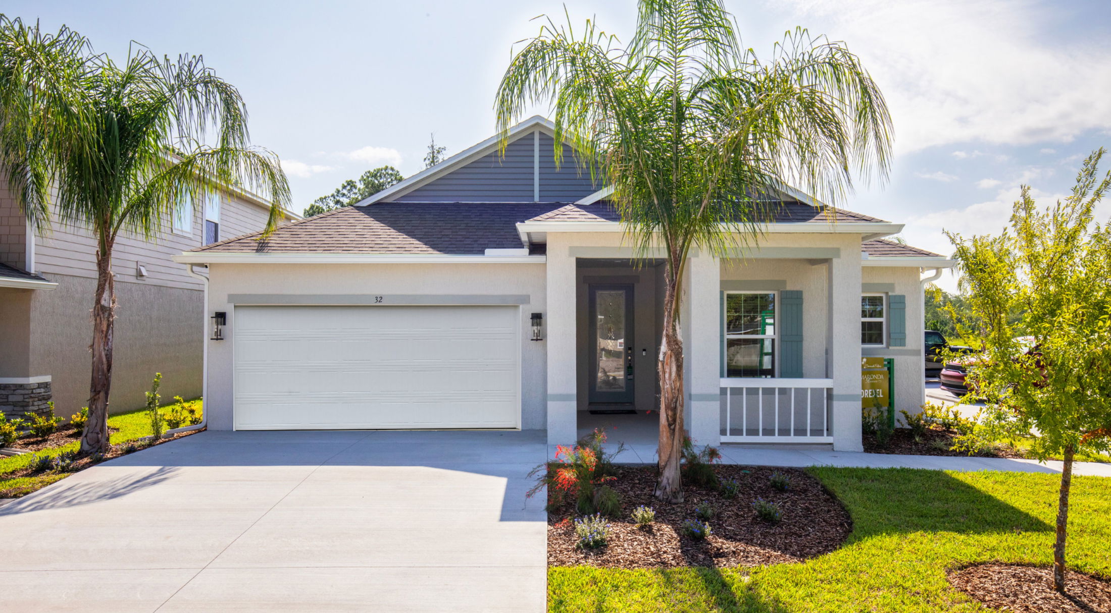 Front view of a modern suburban home with a two-car garage, manicured lawn, and palm trees, under a clear blue sky.