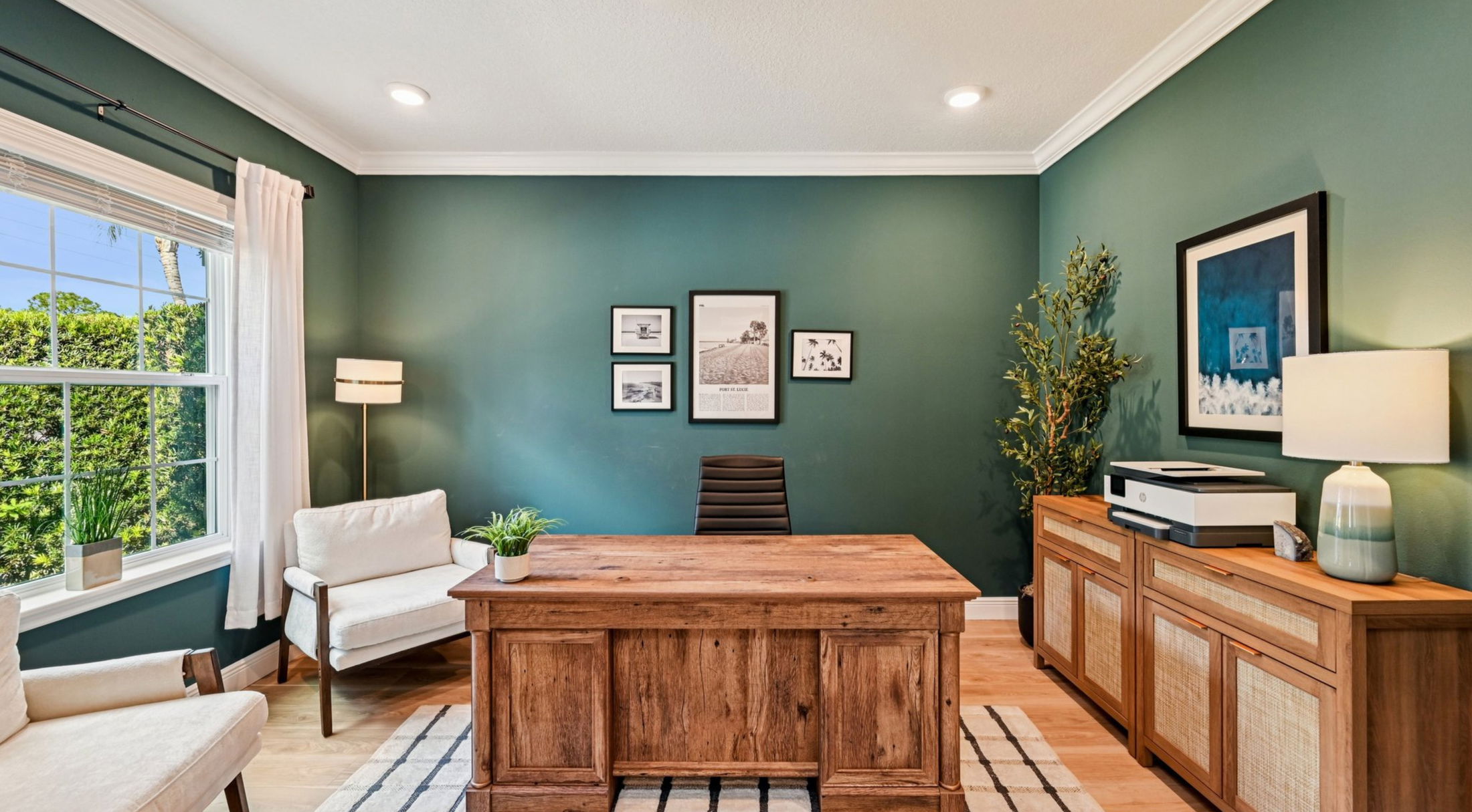 Modern home office with green walls, a wooden desk, white armchairs, and framed wall art.
