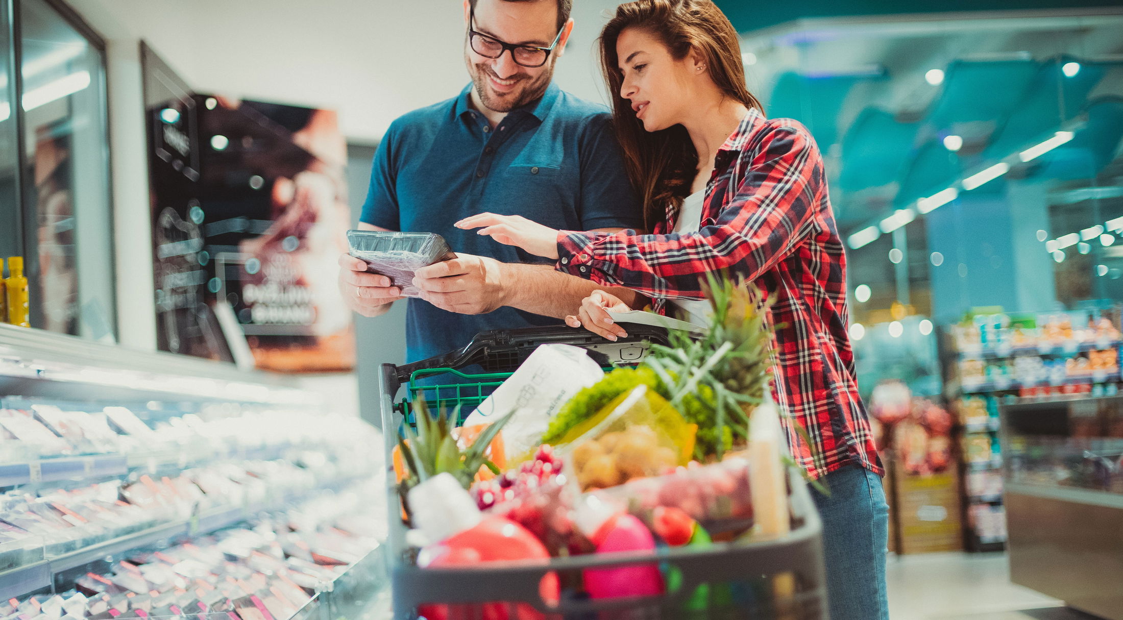 A couple browsing packaged meat in a grocery store aisle while pushing a shopping cart filled with fresh produce.