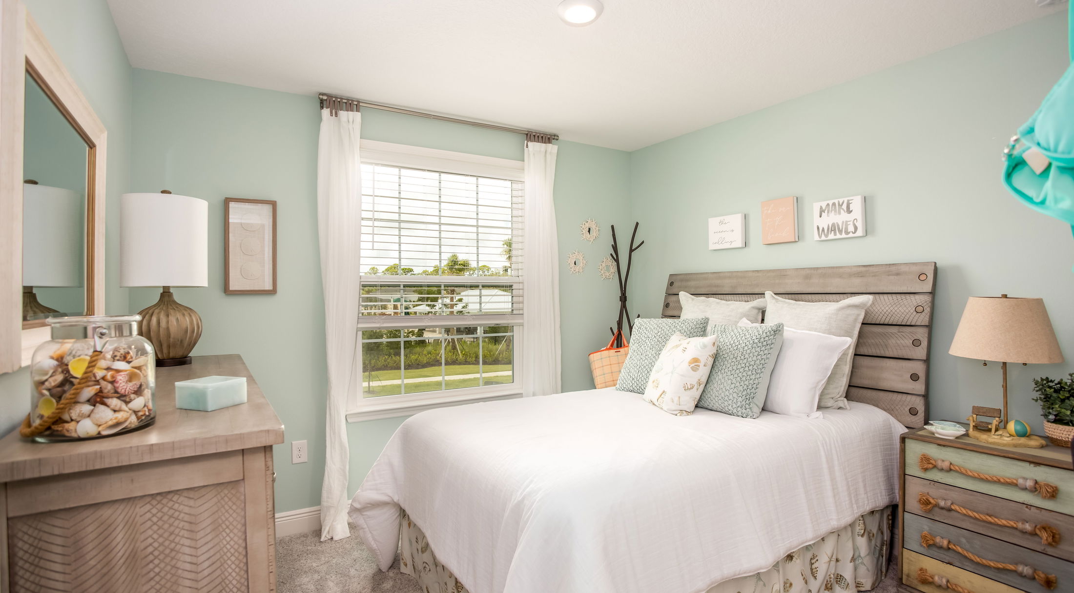 Coastal-themed bedroom with mint green walls, white bedding, nautical decor, seashell accents, and bright natural light from a large window.