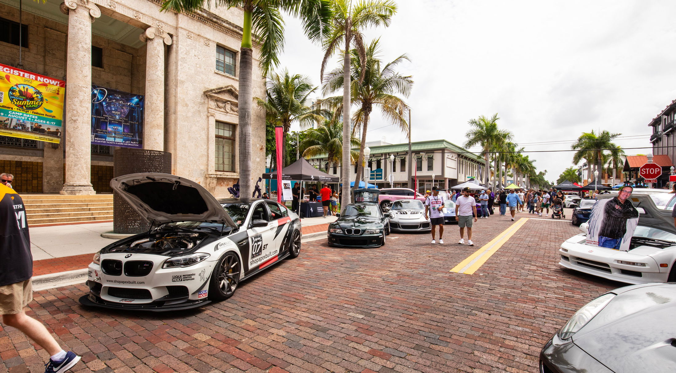 Outdoor car show with various modified cars displayed on a brick street lined with palm trees and historic buildings.