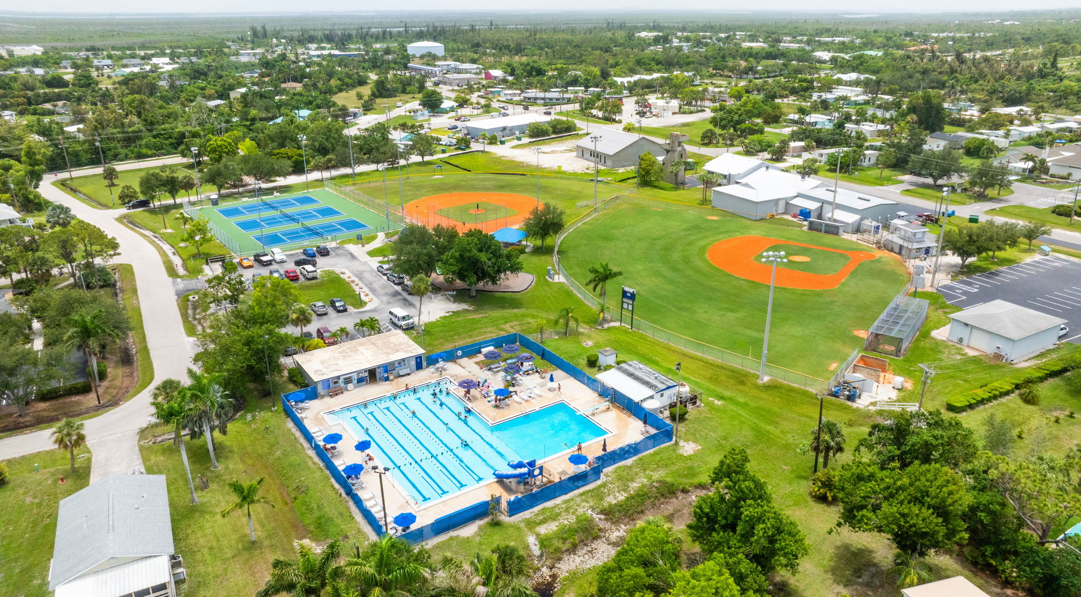 Aerial view of a sports complex featuring a swimming pool, tennis courts, and a baseball field surrounded by greenery and residential areas.