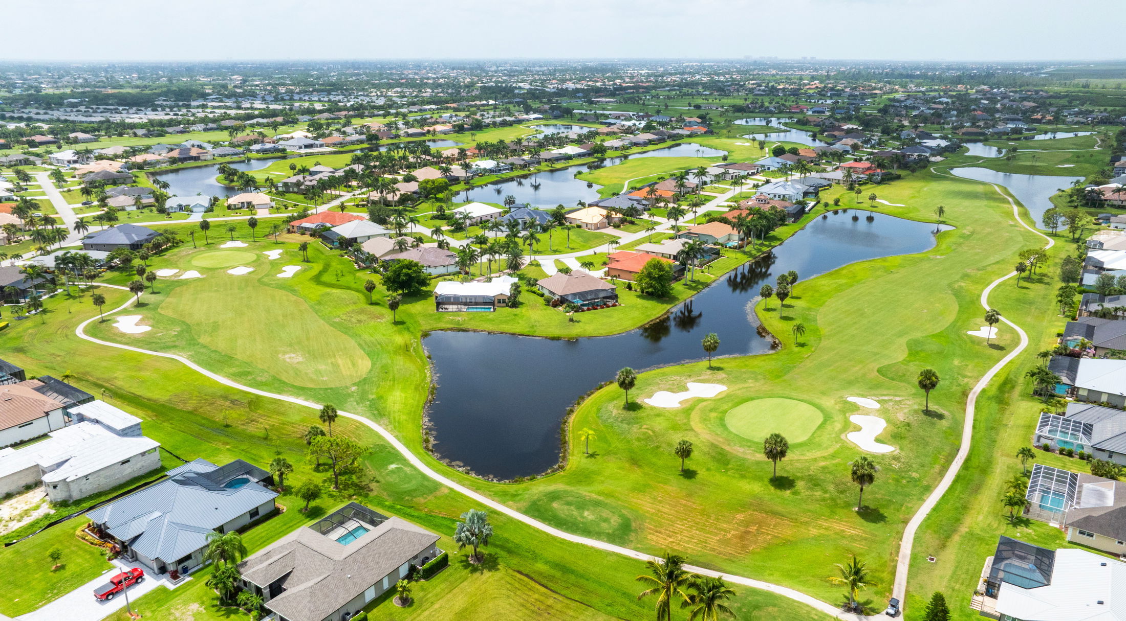 Aerial view of a lush, green golf course with sand bunkers and surrounding residential area, featuring houses with pools and palm trees.