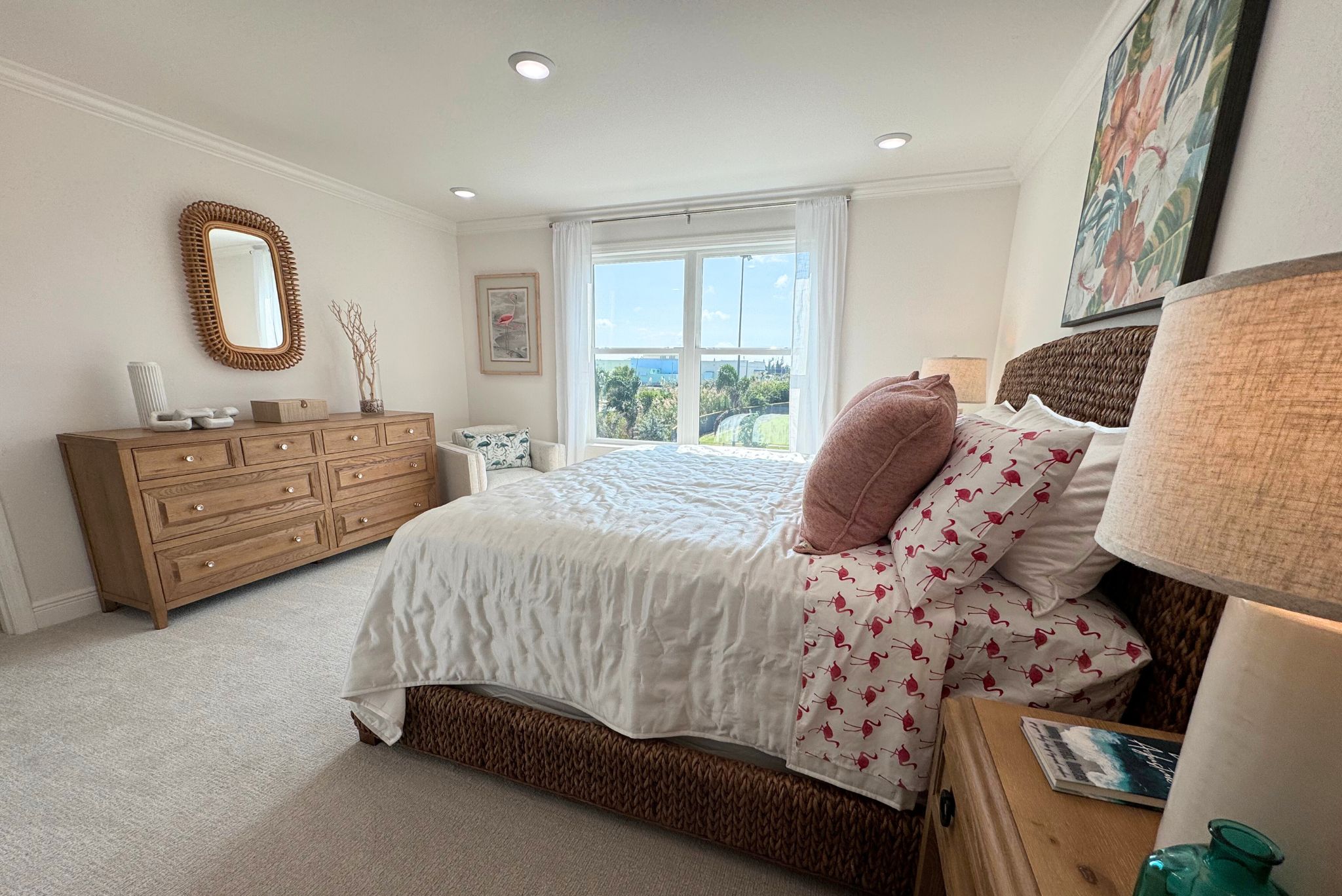 Cozy bedroom interior with a wicker bed frame, pink flamingo bedding, wooden dresser, and large window with a scenic view.