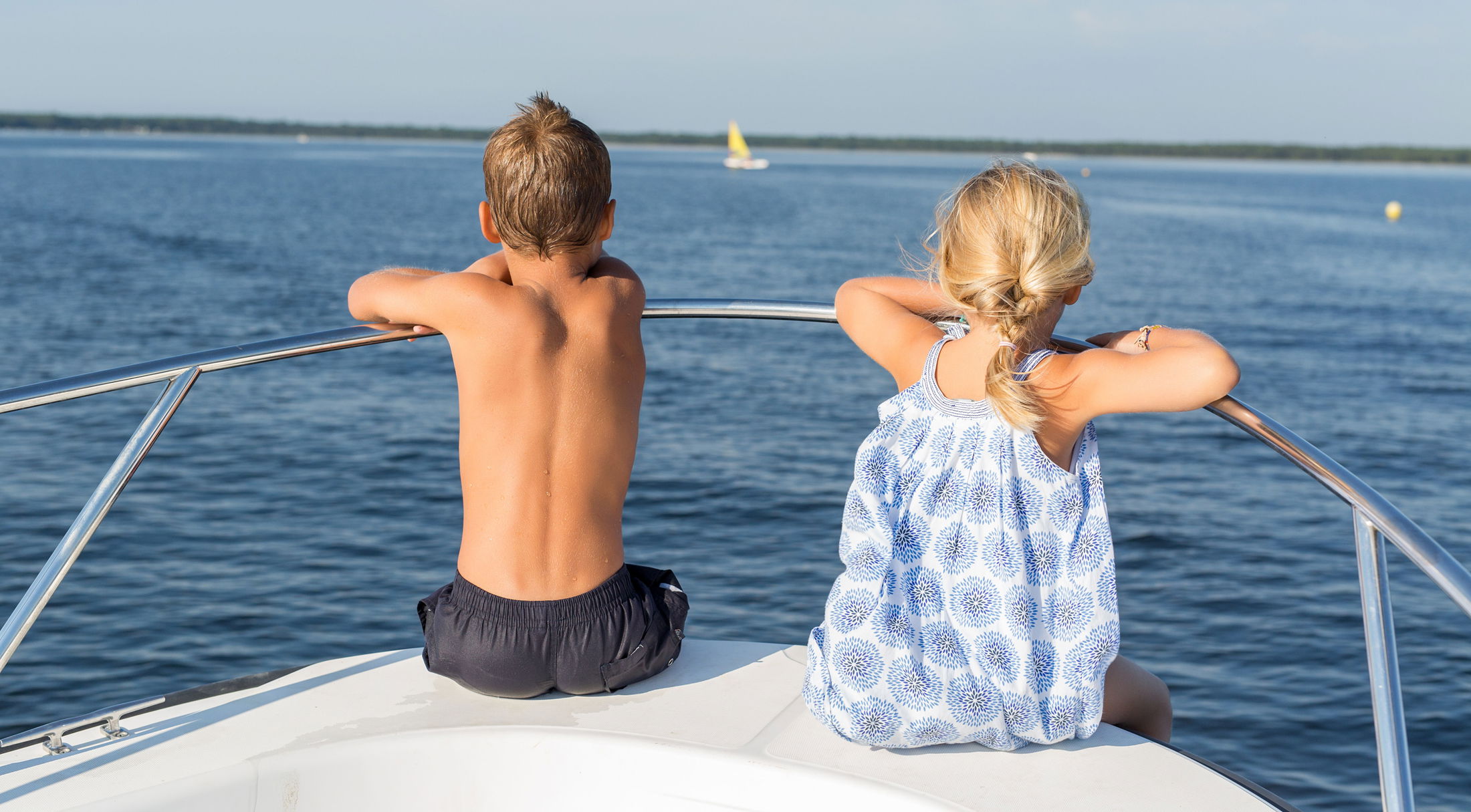 Two children enjoying a sunny day on a boat, overlooking a calm blue lake with a distant yellow sailboat.