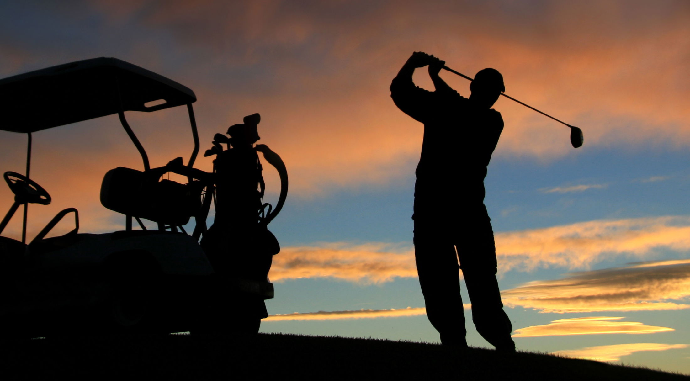 Silhouette of a golfer swinging a club near a golf cart against a vibrant sunset sky.