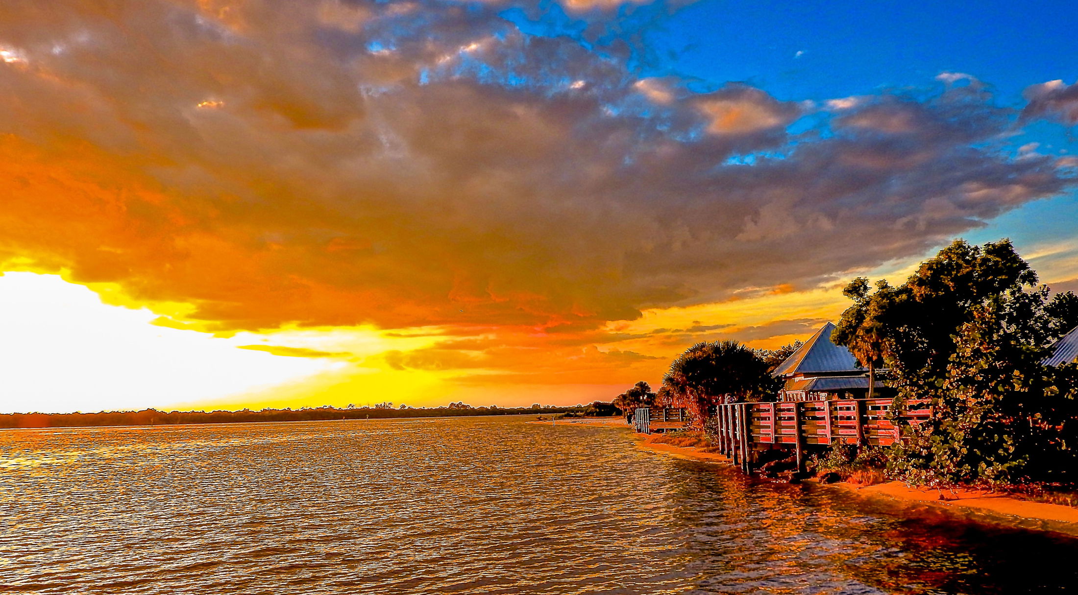 Sunset over a serene lake with vibrant clouds and rustic waterfront cottages surrounded by lush trees.