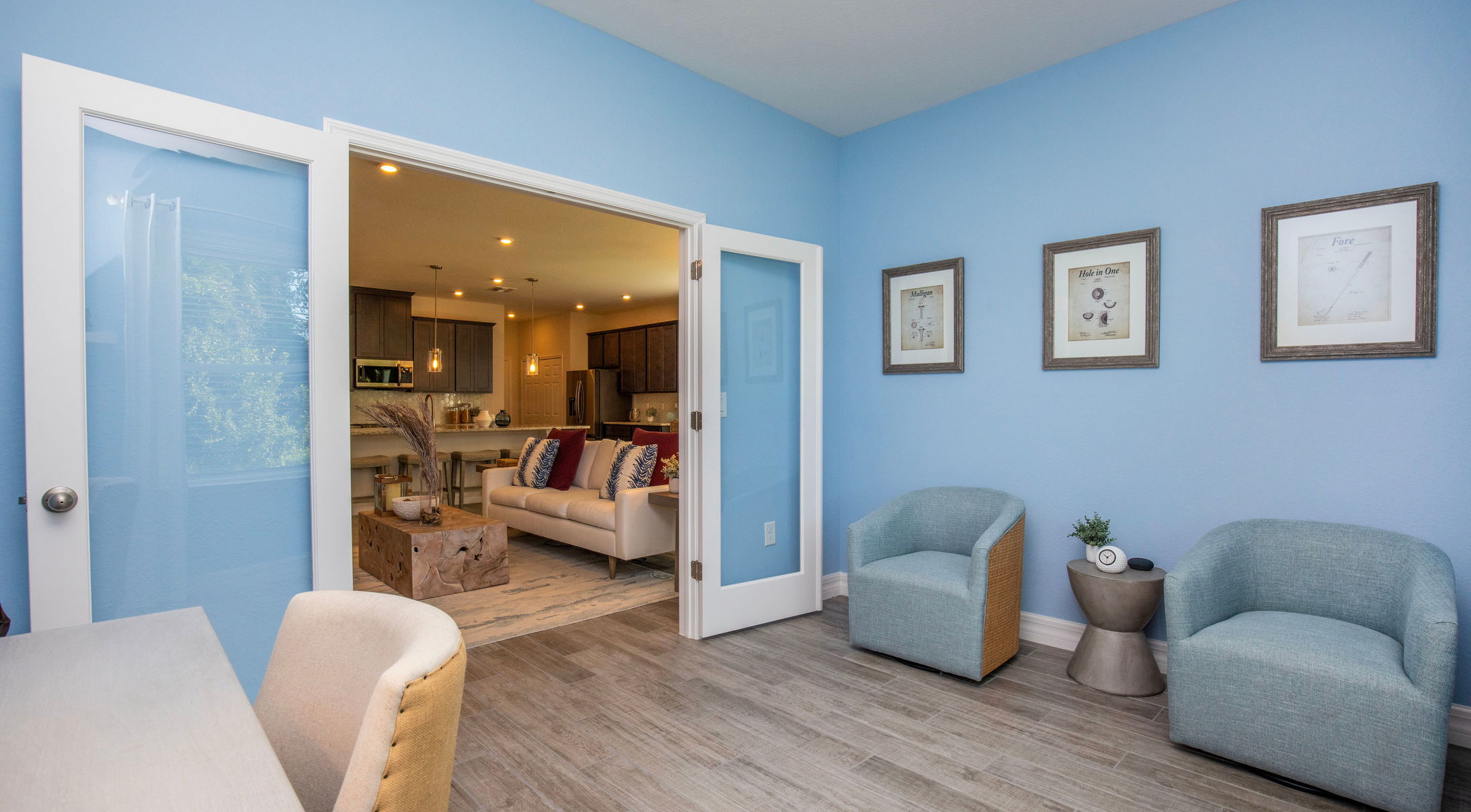 Cozy modern living room with blue walls, framed artwork, and open glass doors leading to an inviting kitchen area.