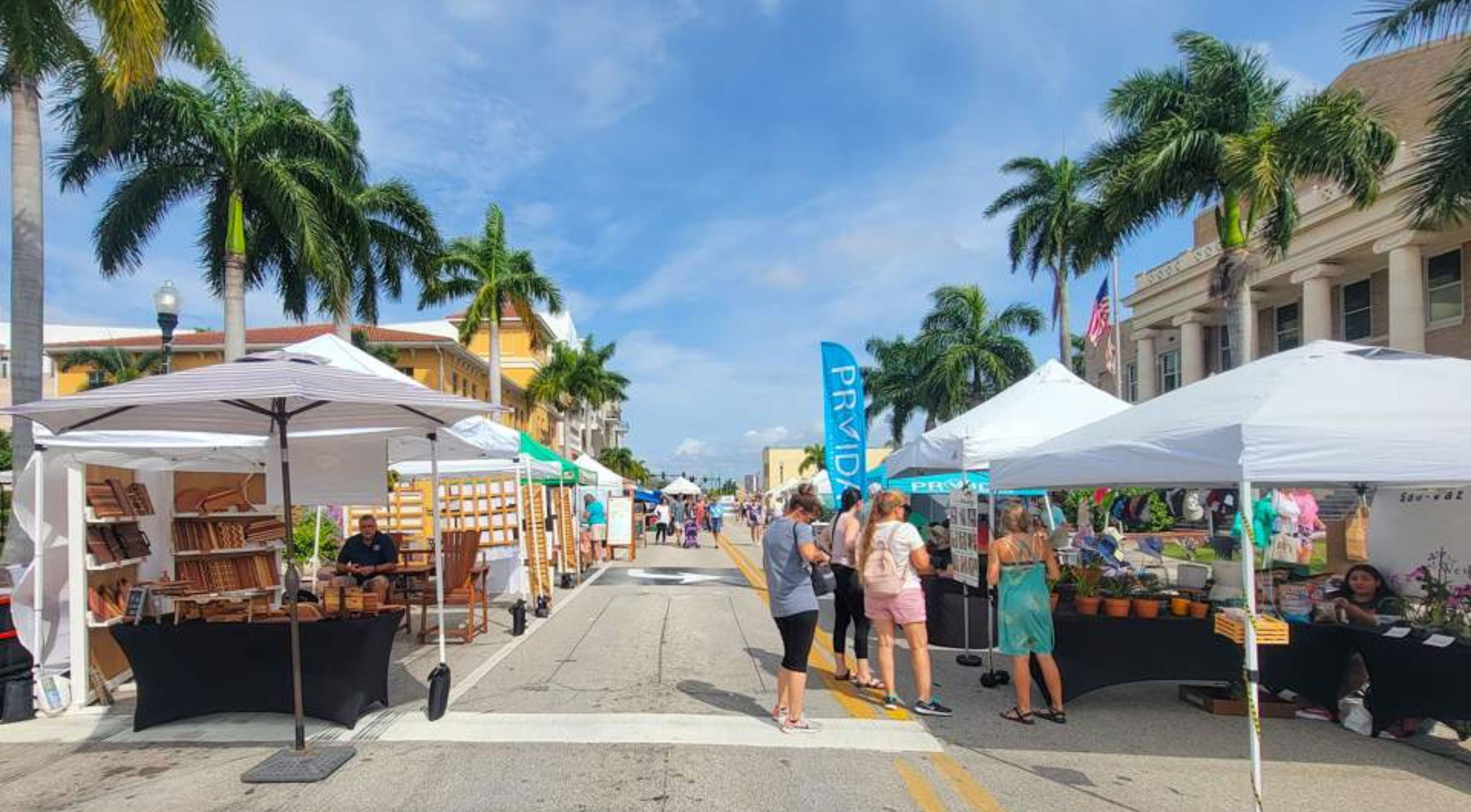 Colorful outdoor market with vendors and shoppers under tents on a sunny day surrounded by palm trees.