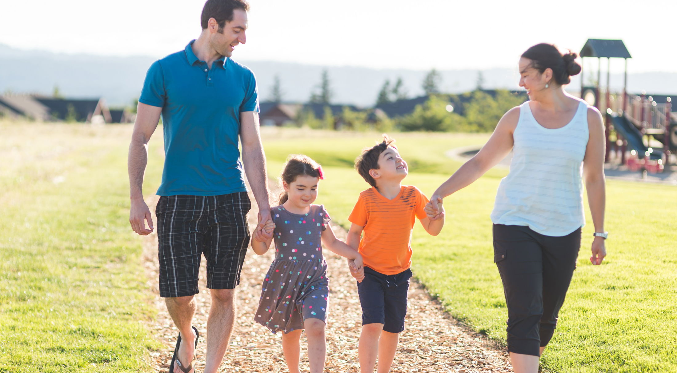 A smiling family of four enjoys a sunny walk together on a park path.