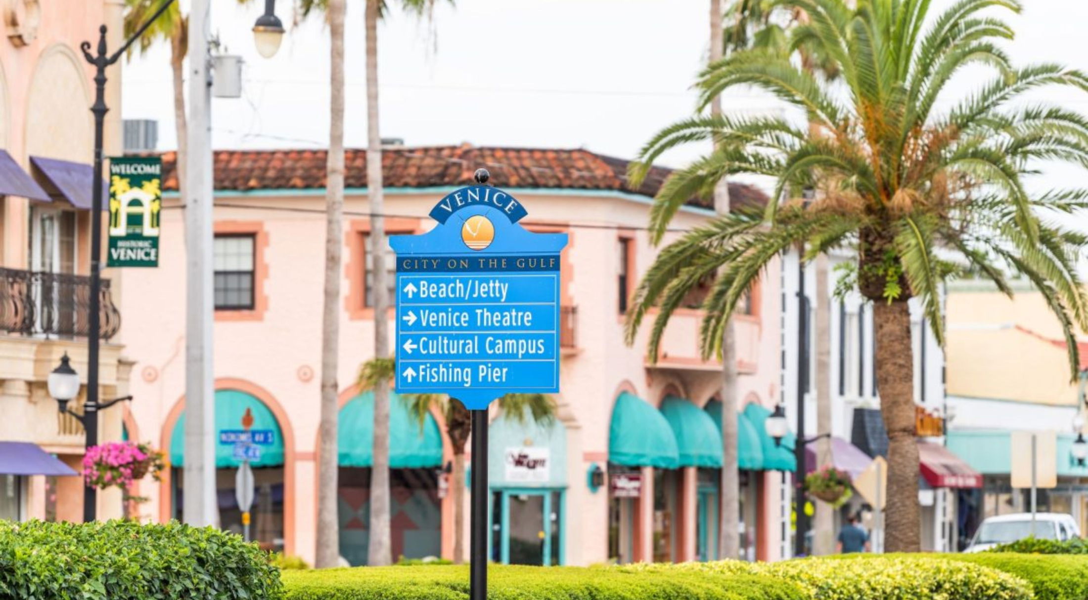 Blue directional sign in Venice, Florida guiding to beach, theater, cultural campus, and fishing pier, with palm trees and pastel buildings in the background.