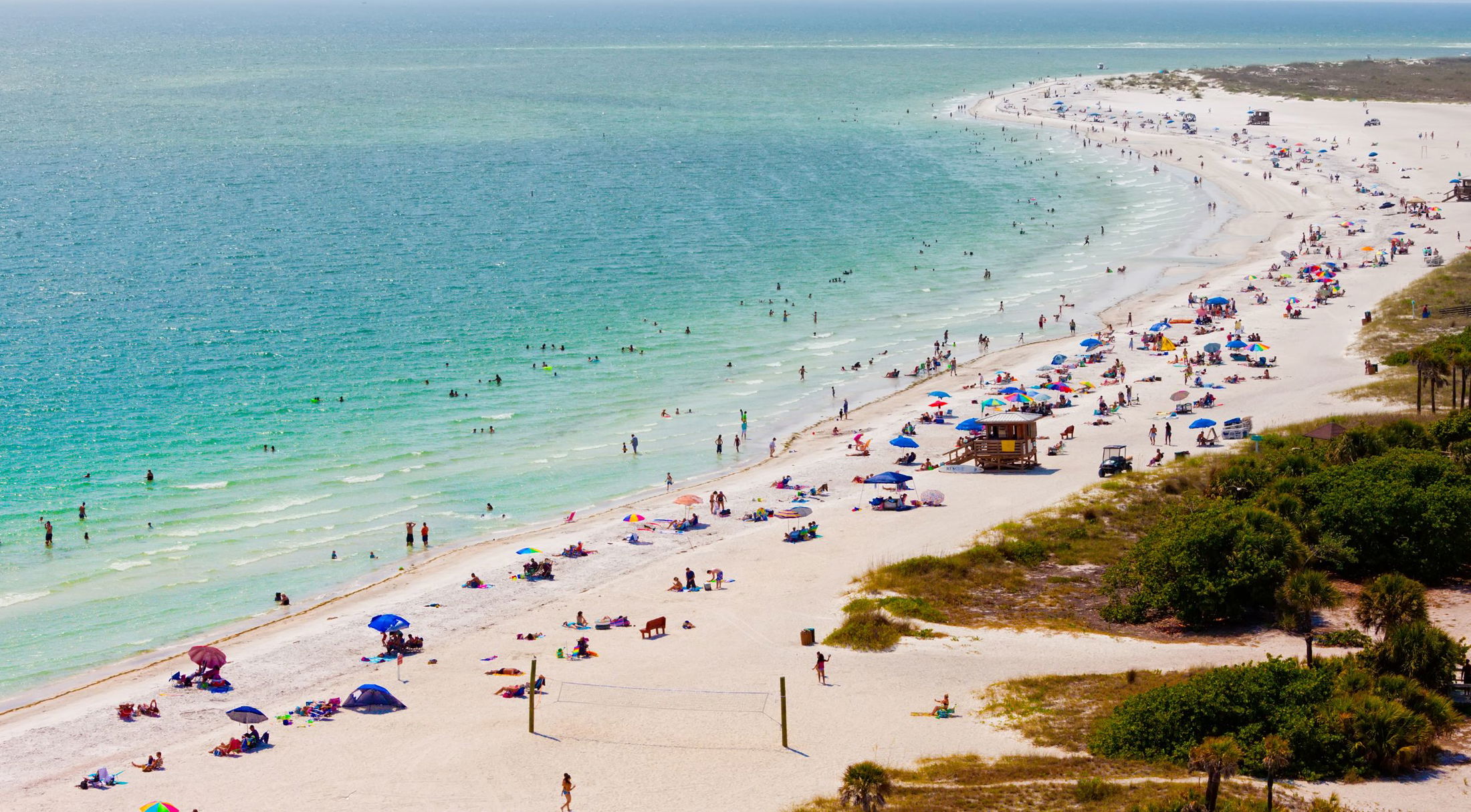 Aerial view of a bustling white sand beach with colorful umbrellas and people swimming in clear blue ocean water.