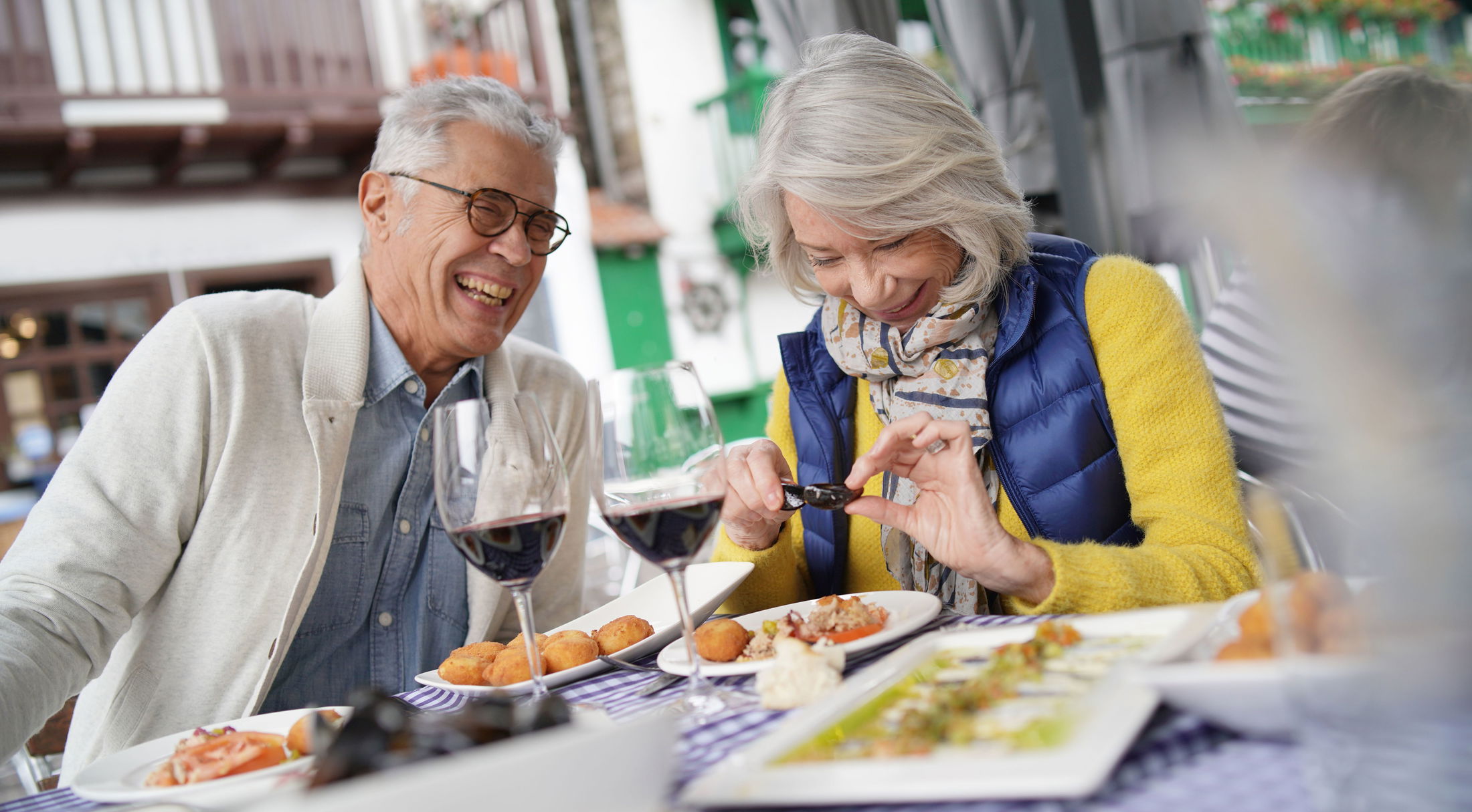 Elderly couple enjoying a meal with wine at an outdoor restaurant in a vibrant, joyful atmosphere.