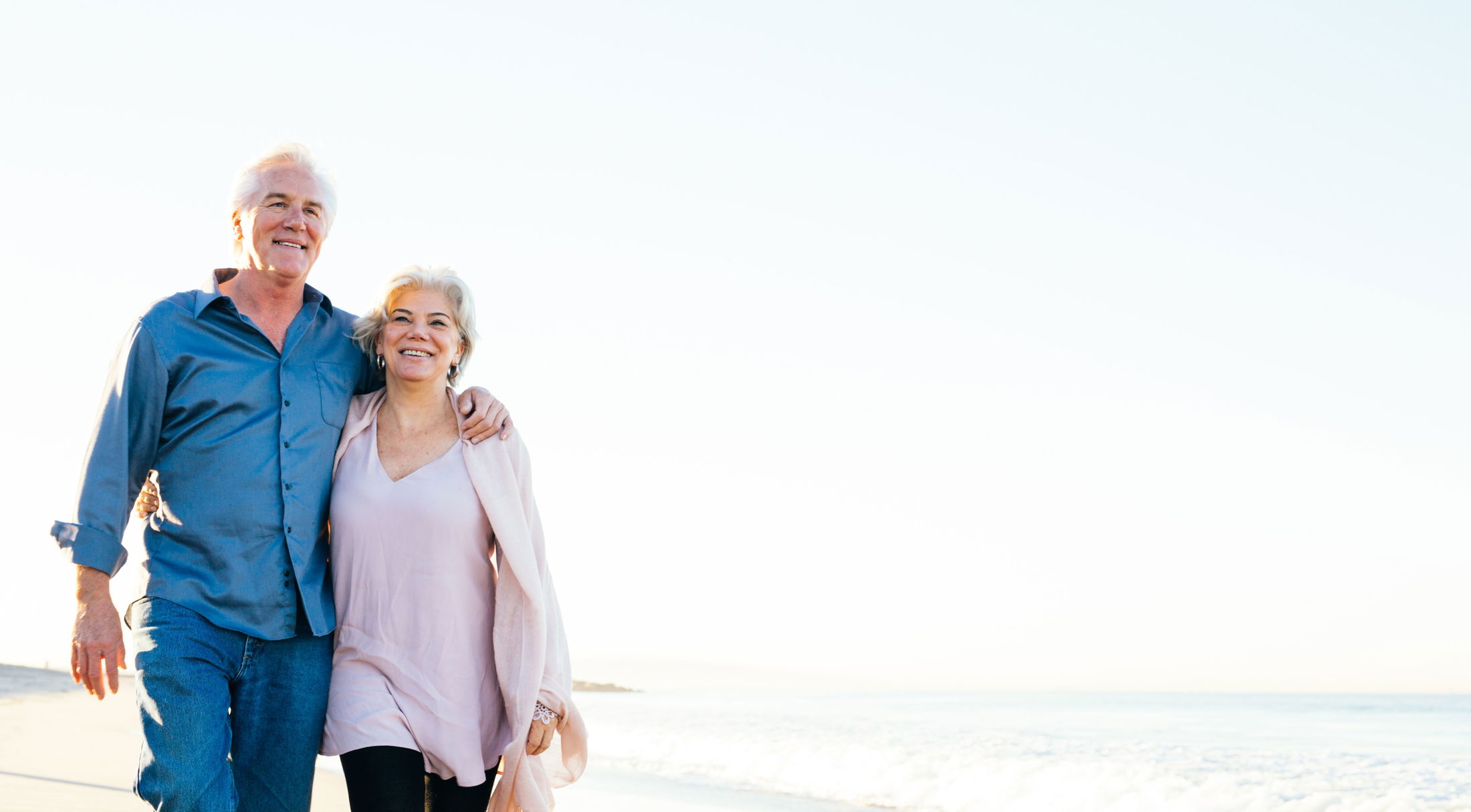 Senior couple happily walking on a sunny beach, embracing and smiling.