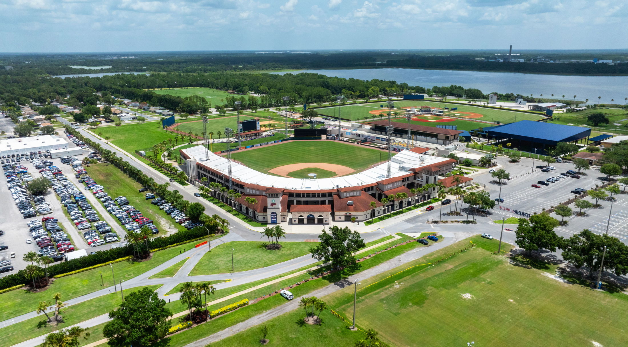 Aerial view of a baseball stadium with surrounding parking lots and lush greenery, situated near a river under a partly cloudy sky.