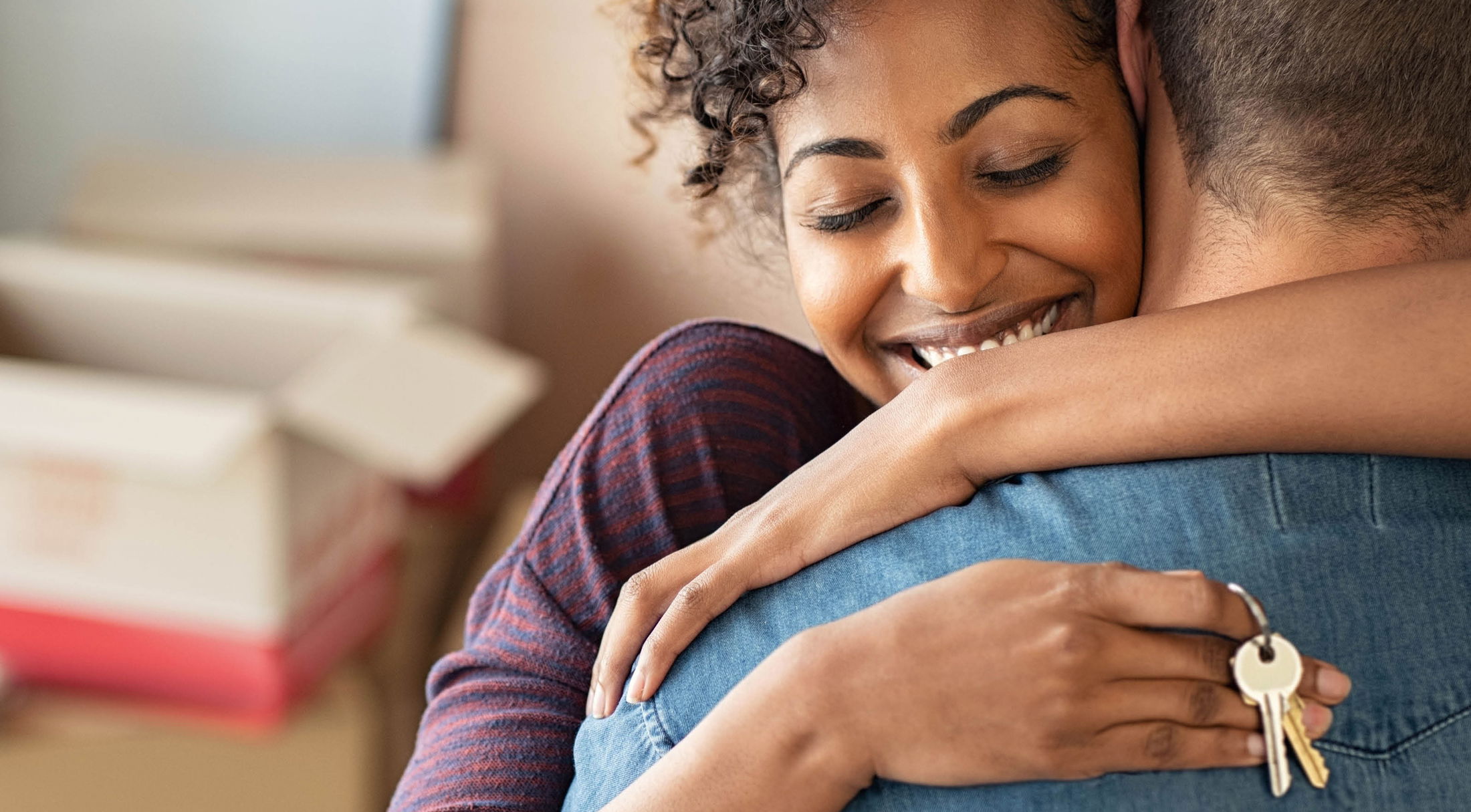 A joyful woman with keys in hand embraces a man amidst moving boxes, symbolizing a new home or moving day.