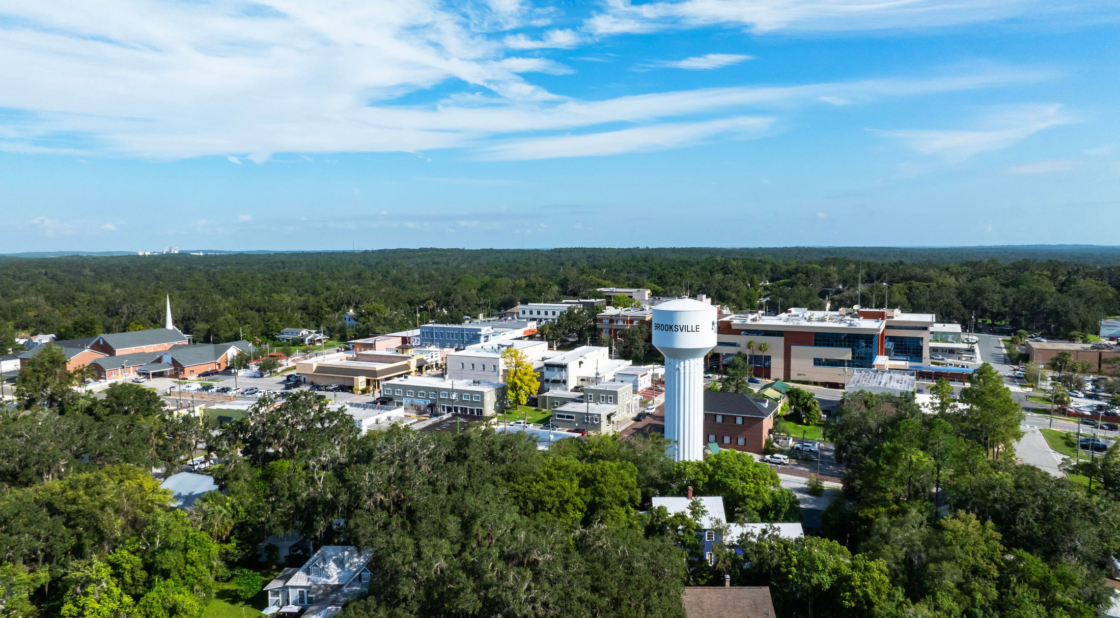 Aerial view of Brooksville, Florida, featuring its prominent water tower surrounded by lush greenery and urban buildings under a clear blue sky.