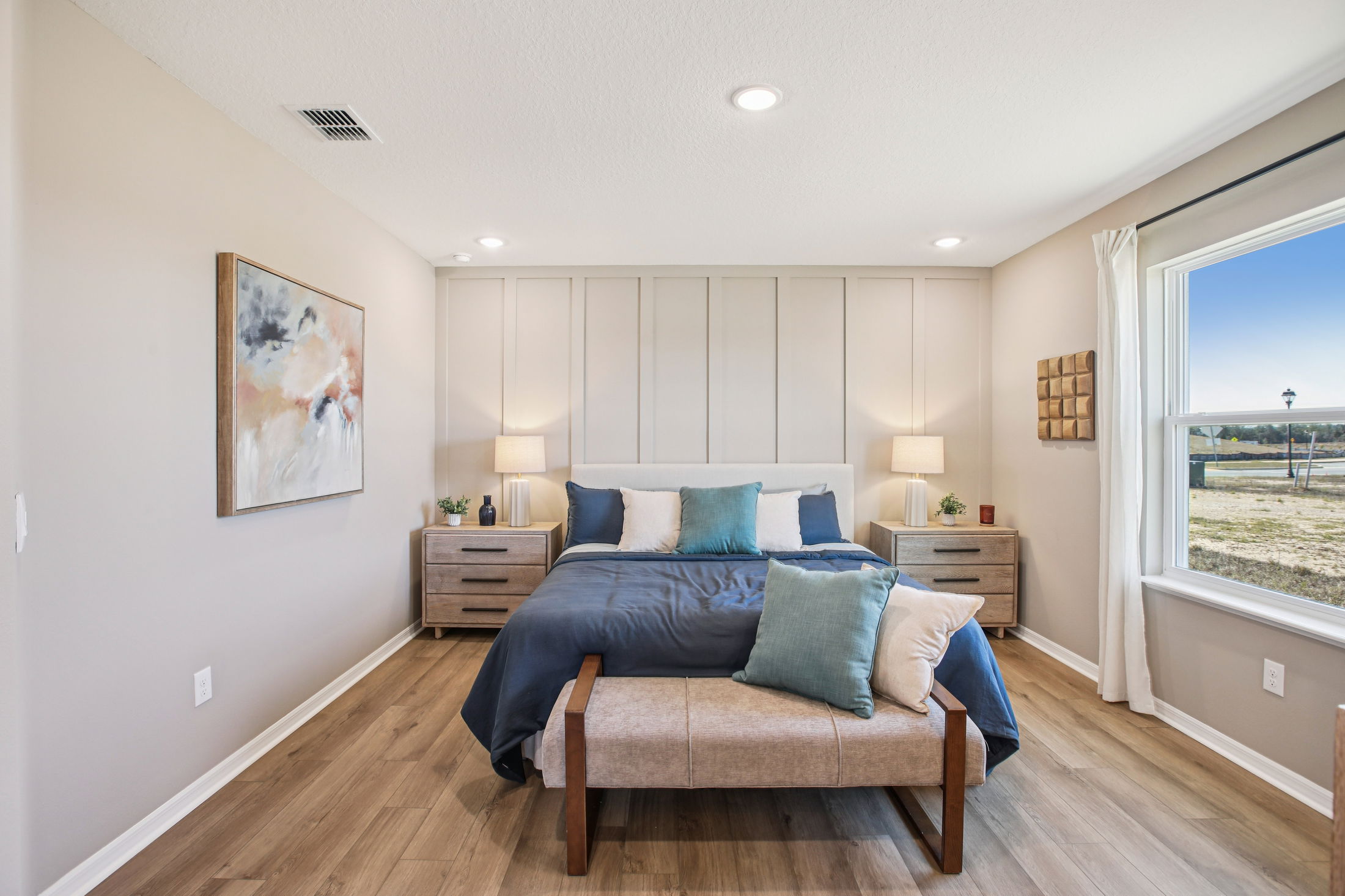 Modern bedroom featuring a plush bed with blue and white pillows, stylish nightstands, and large windows illuminating the space.