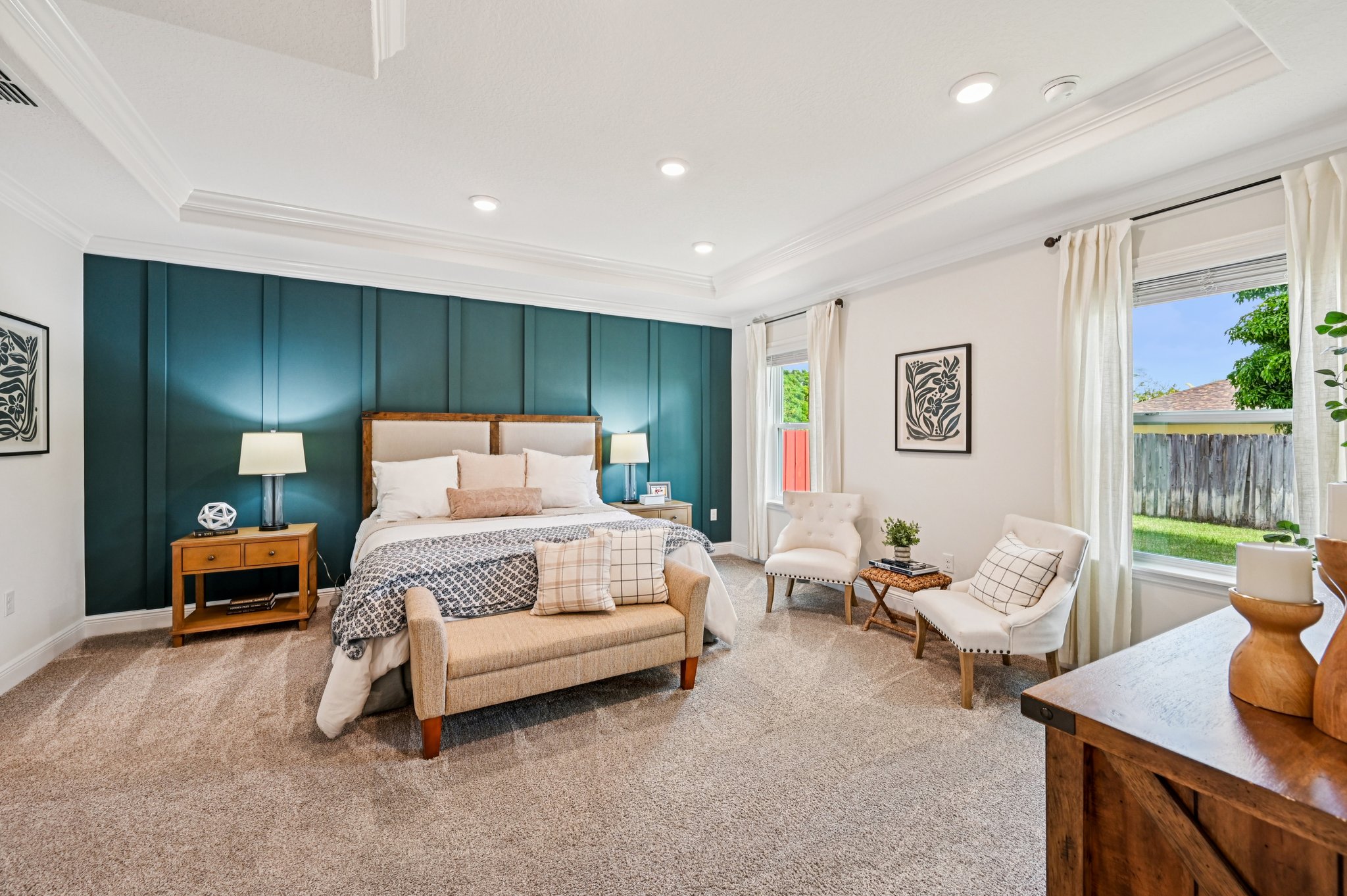 A stylish bedroom featuring a bed with decorative pillows, two bedside lamps, an armchair, and large windows with sheer curtains.