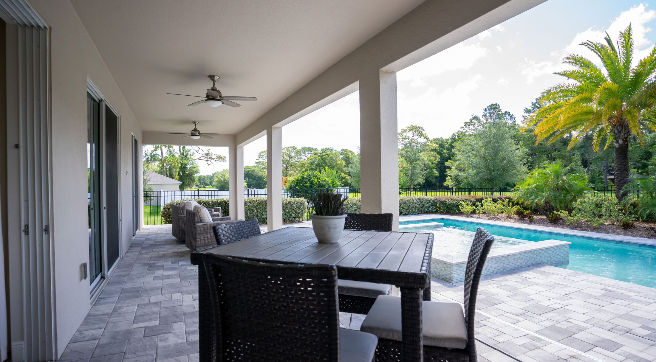 Covered patio with wicker furniture overlooking a swimming pool and lush garden landscape.