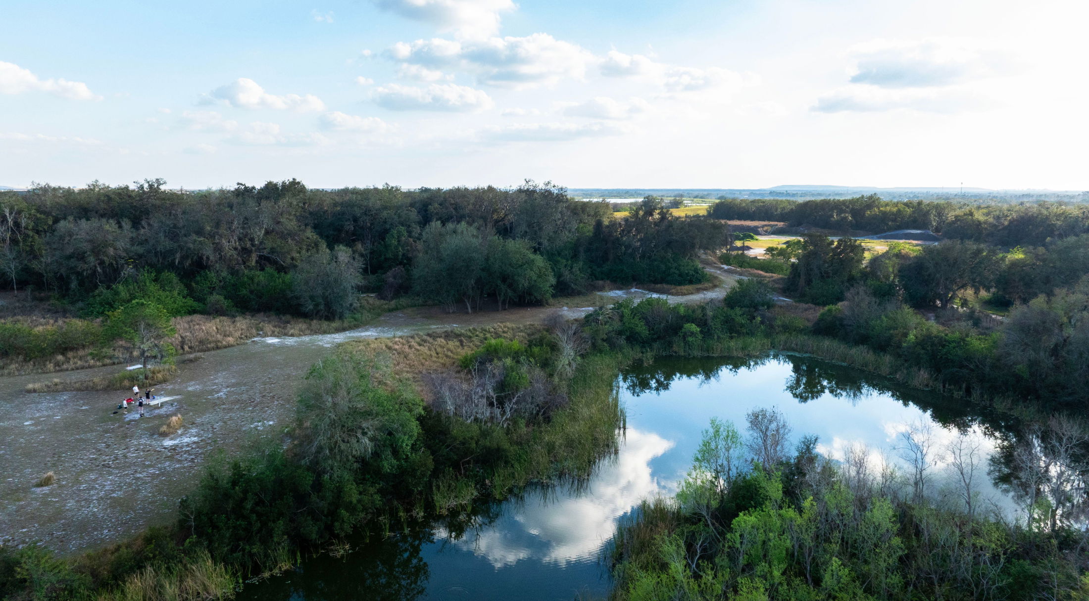 Aerial view of a scenic landscape featuring a tranquil lake surrounded by lush greenery and a group of people gathering on the riverside.