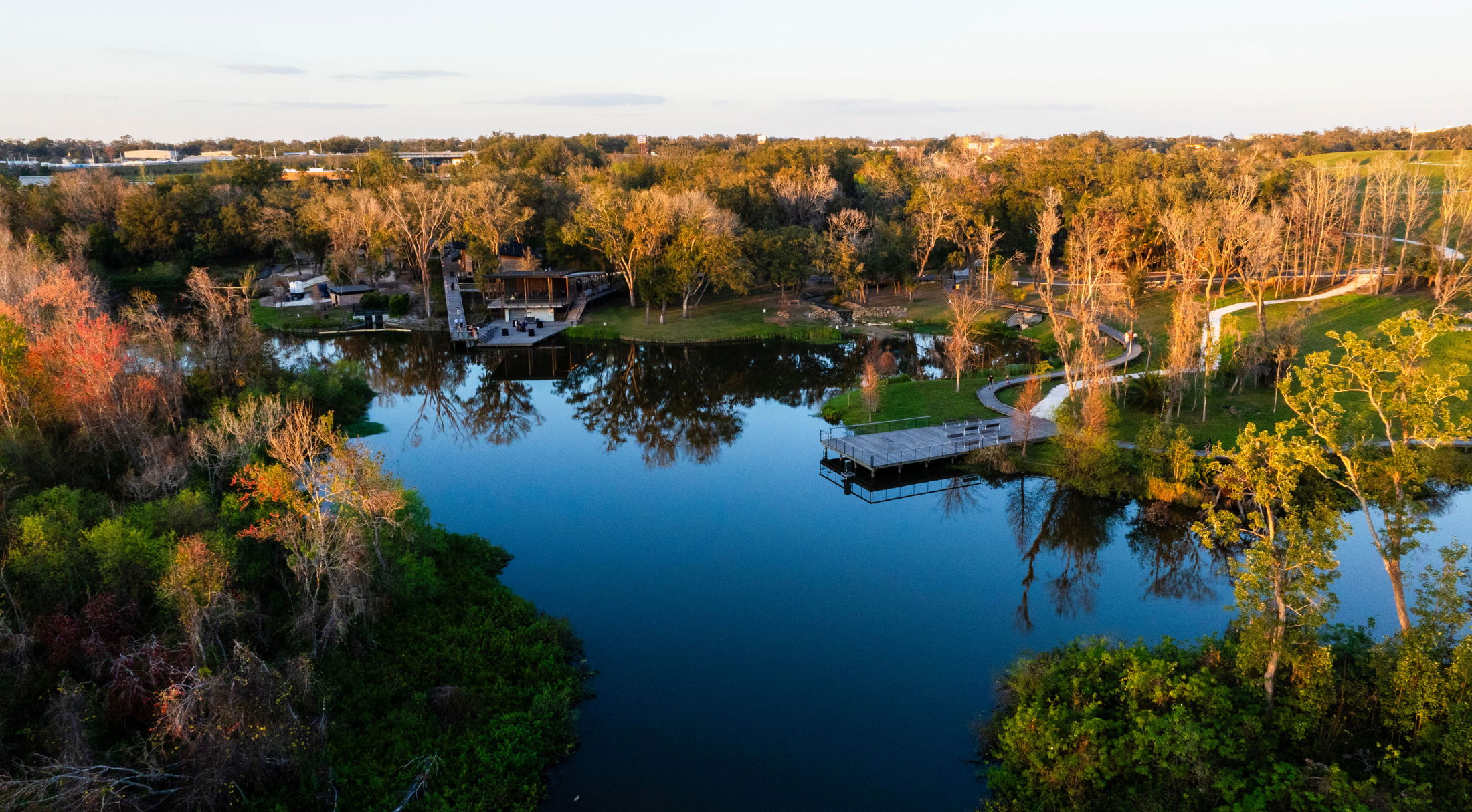 Aerial view of a scenic park landscape with a reflective lake, surrounded by trees and walking paths at sunset.