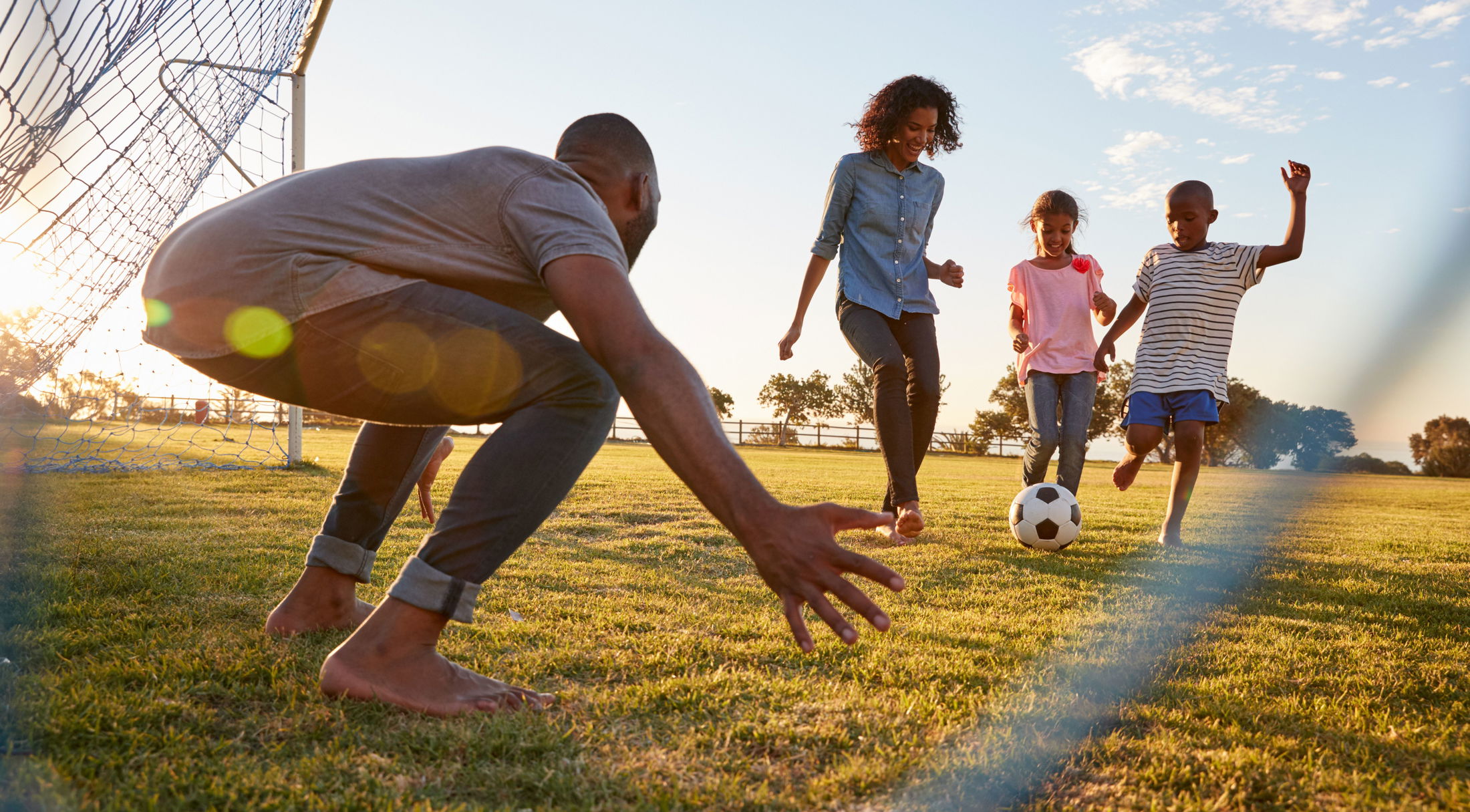 A family enjoying a game of soccer in a sunny park, with children kicking the ball towards a crouching adult goalie near the net.