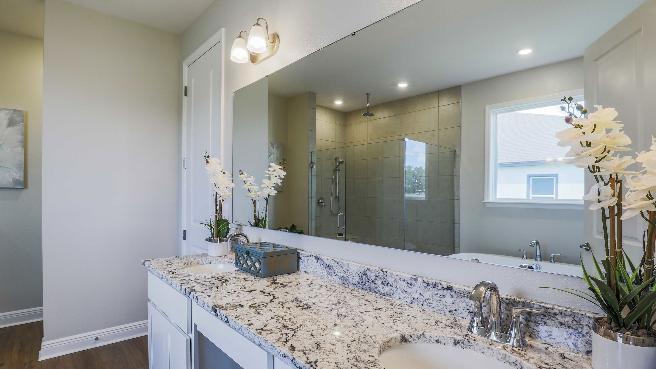 Modern bathroom with a granite countertop, chrome fixtures, and a large mirror reflecting a tiled shower area and white orchid decorations.
