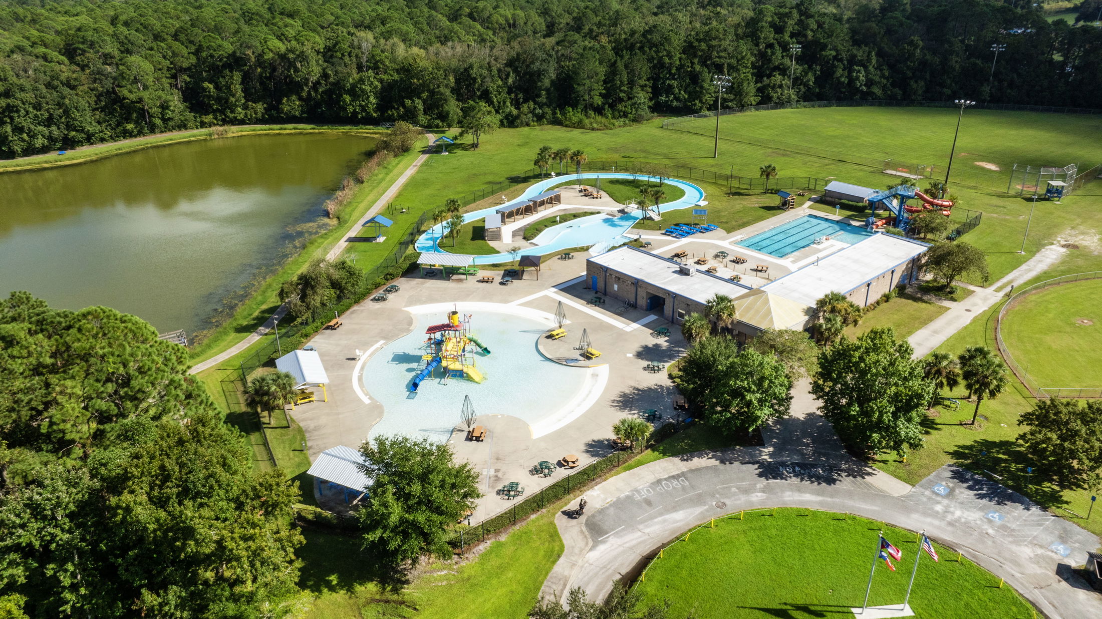 Aerial view of a vibrant outdoor water park with swimming pools, water slides, and a lazy river, surrounded by lush greenery and a pond.