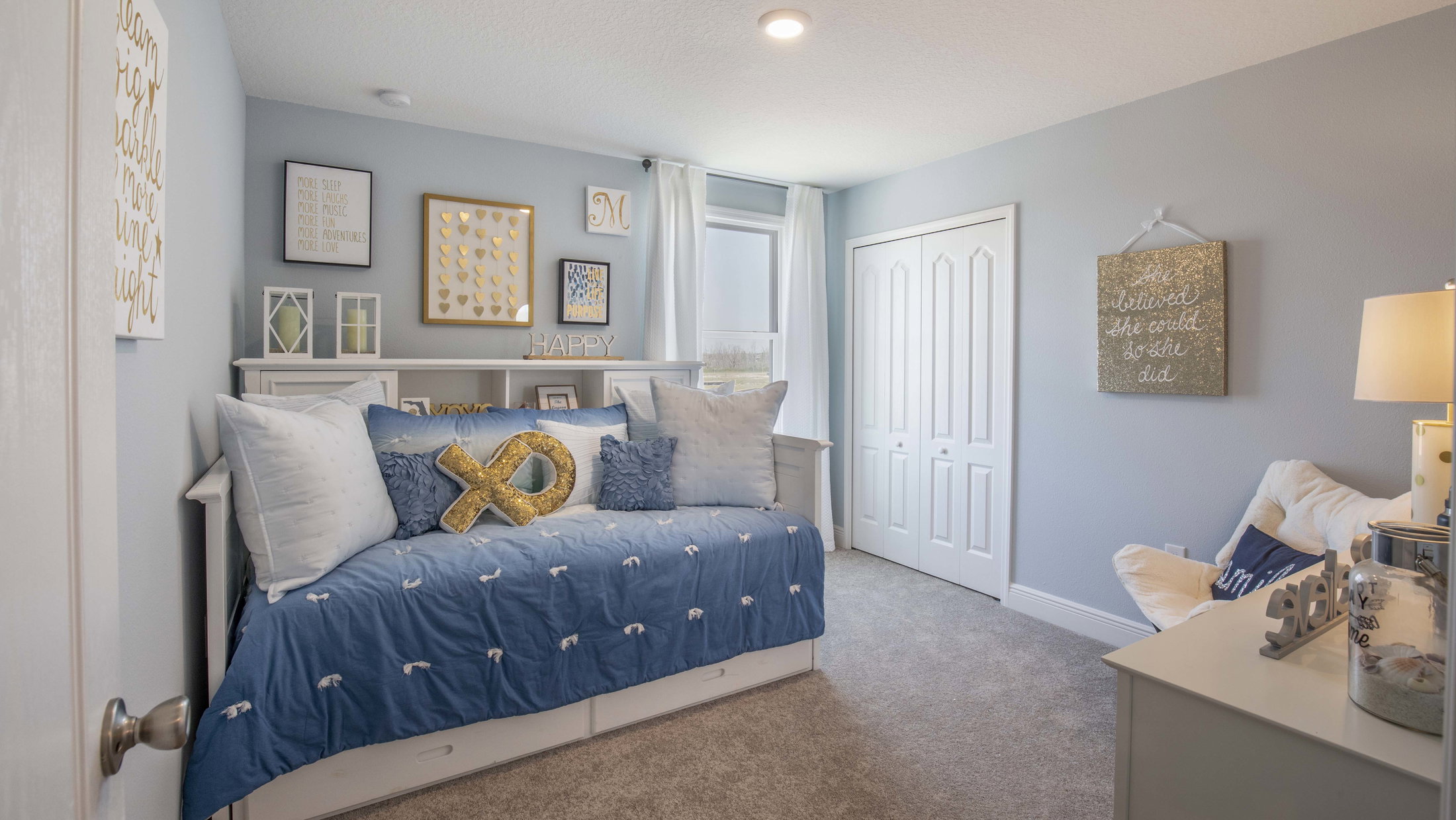 A cozy bedroom featuring a blue daybed with decorative pillows, light blue walls, and framed art on the walls.