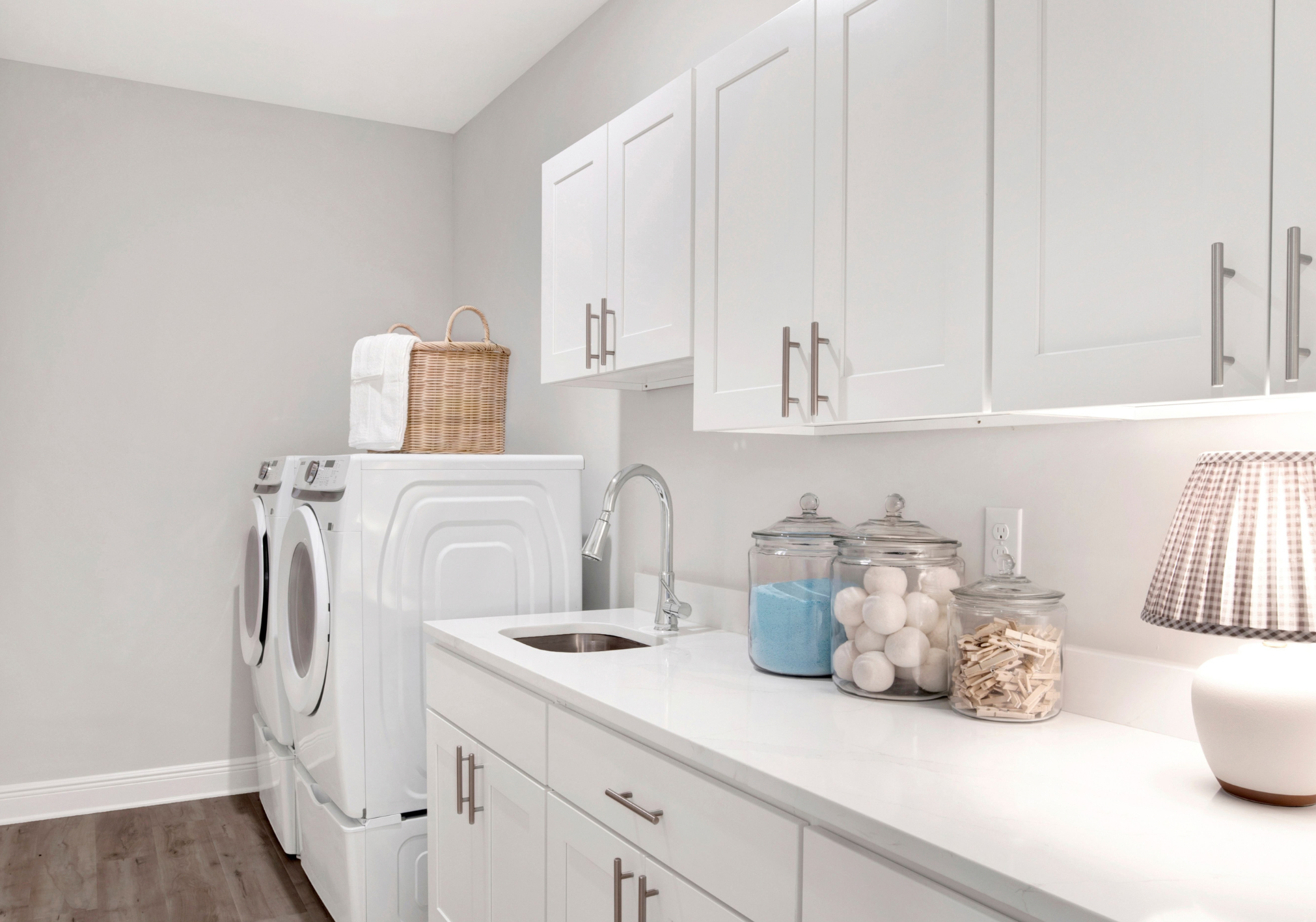 A modern laundry room featuring white cabinets, a stacked washer and dryer, a sink, and decorative jars with laundry supplies.