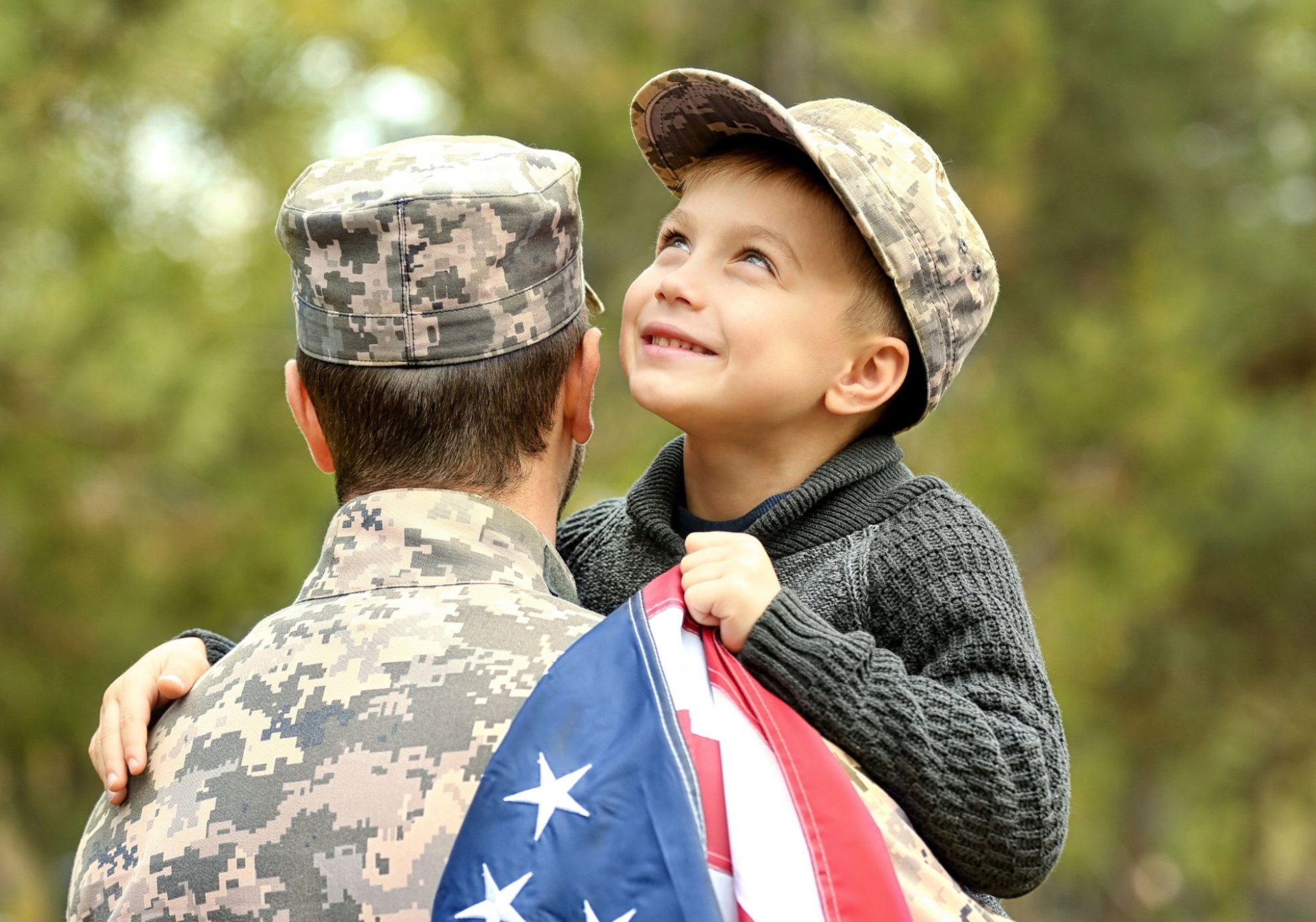 A young boy smiles while being held by a soldier, wrapped in an American flag.
