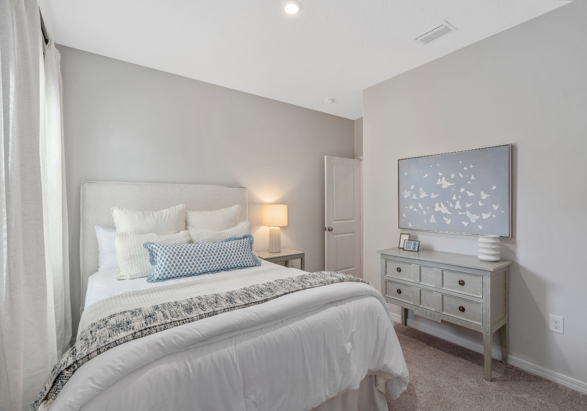 Cozy bedroom featuring a bed with white linens, a blue decorative pillow, a nightstand with a lamp, and a wall-mounted picture of birds.
