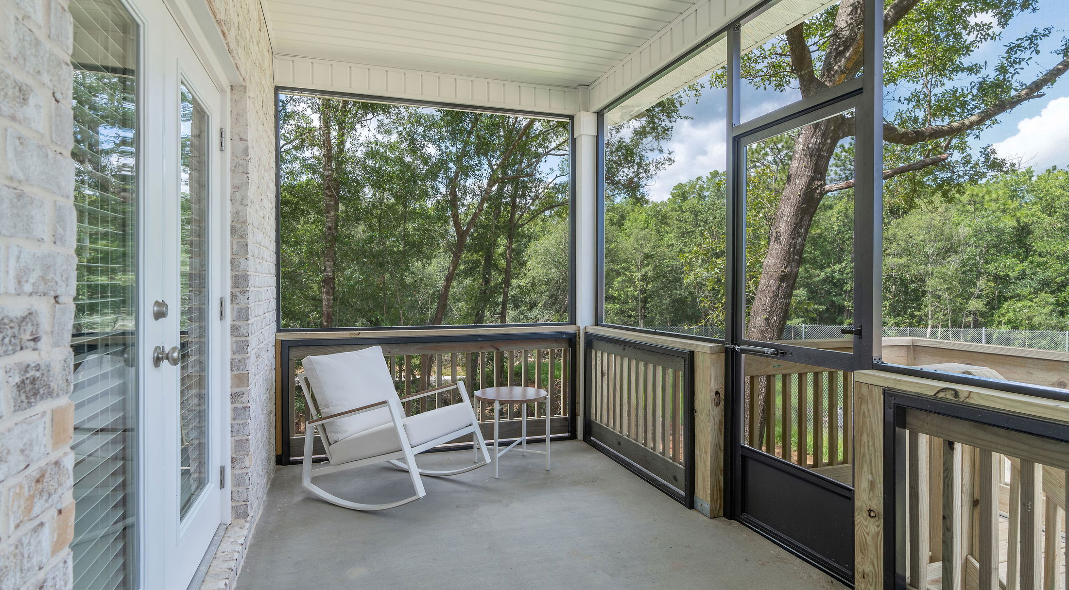 Screened-in porch with a white rocking chair and small table overlooking a lush green forest.