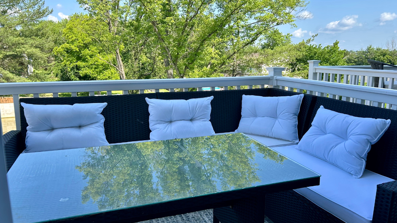 Outdoor patio with white cushioned wicker furniture and glass table on a sunny deck with greenery in the background.