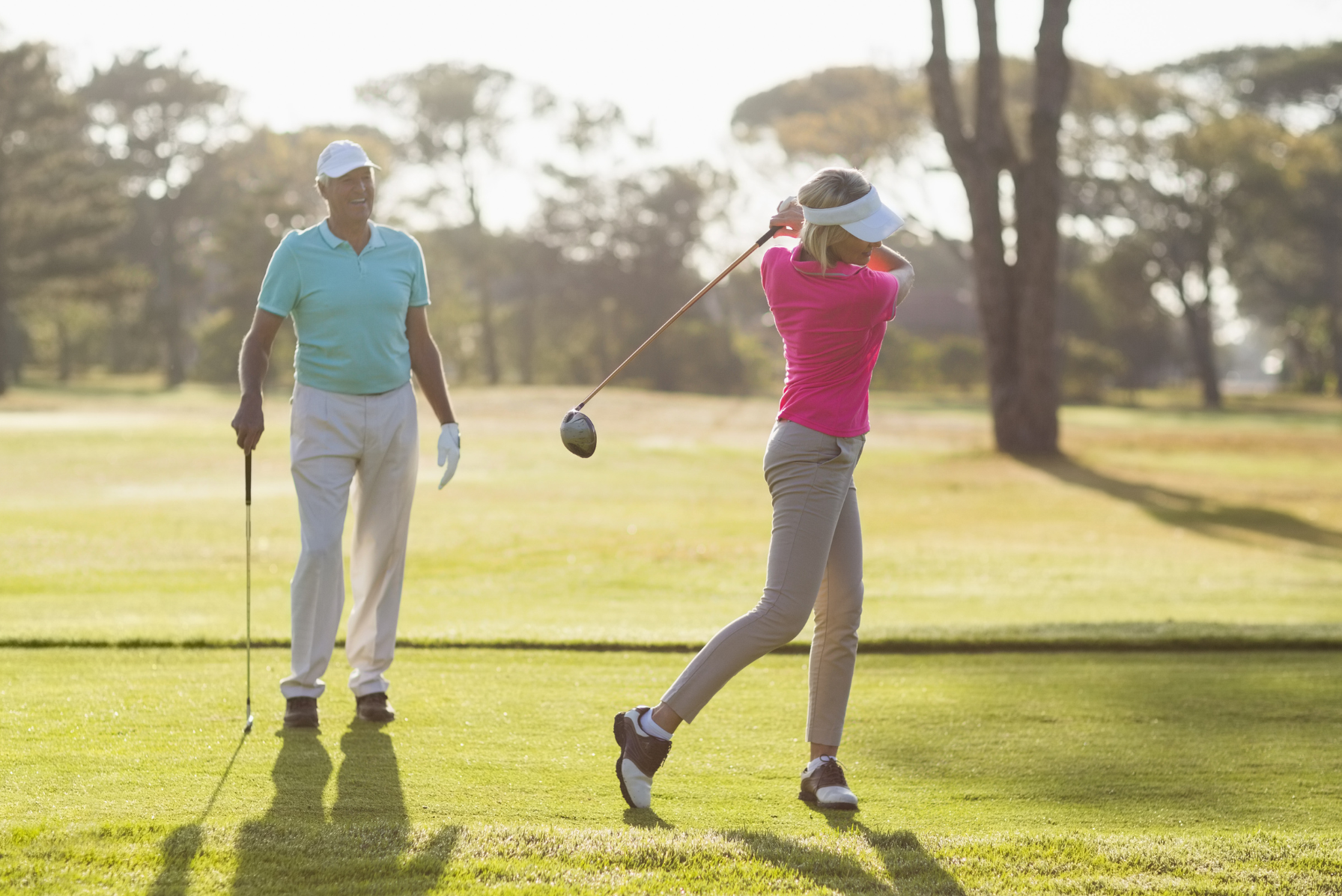 A woman in a pink shirt and visor swings a golf club on a sunny course while a man in a blue shirt watches.