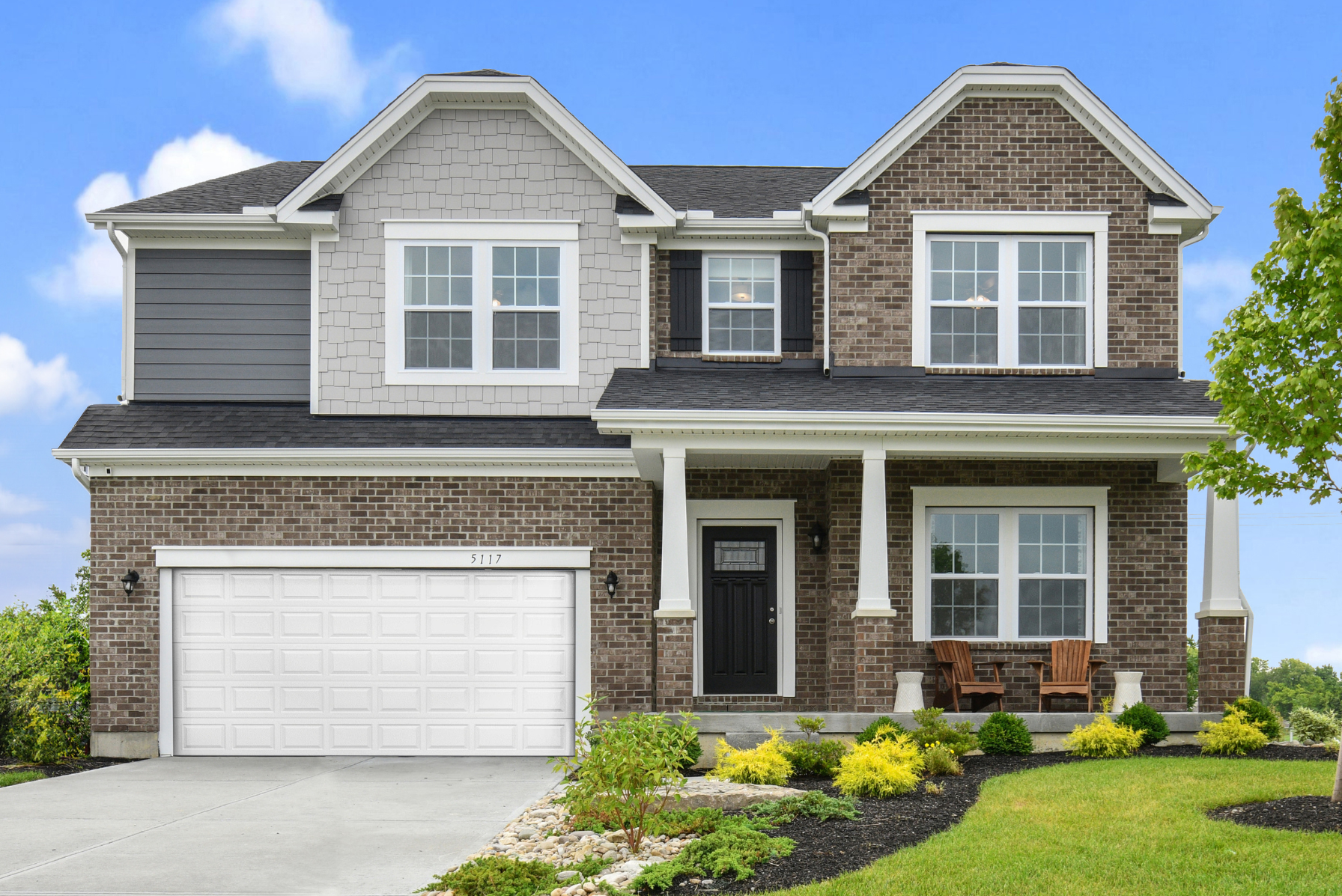 Two-story suburban house with brick facade, landscaped front yard, and a white garage under a clear blue sky.