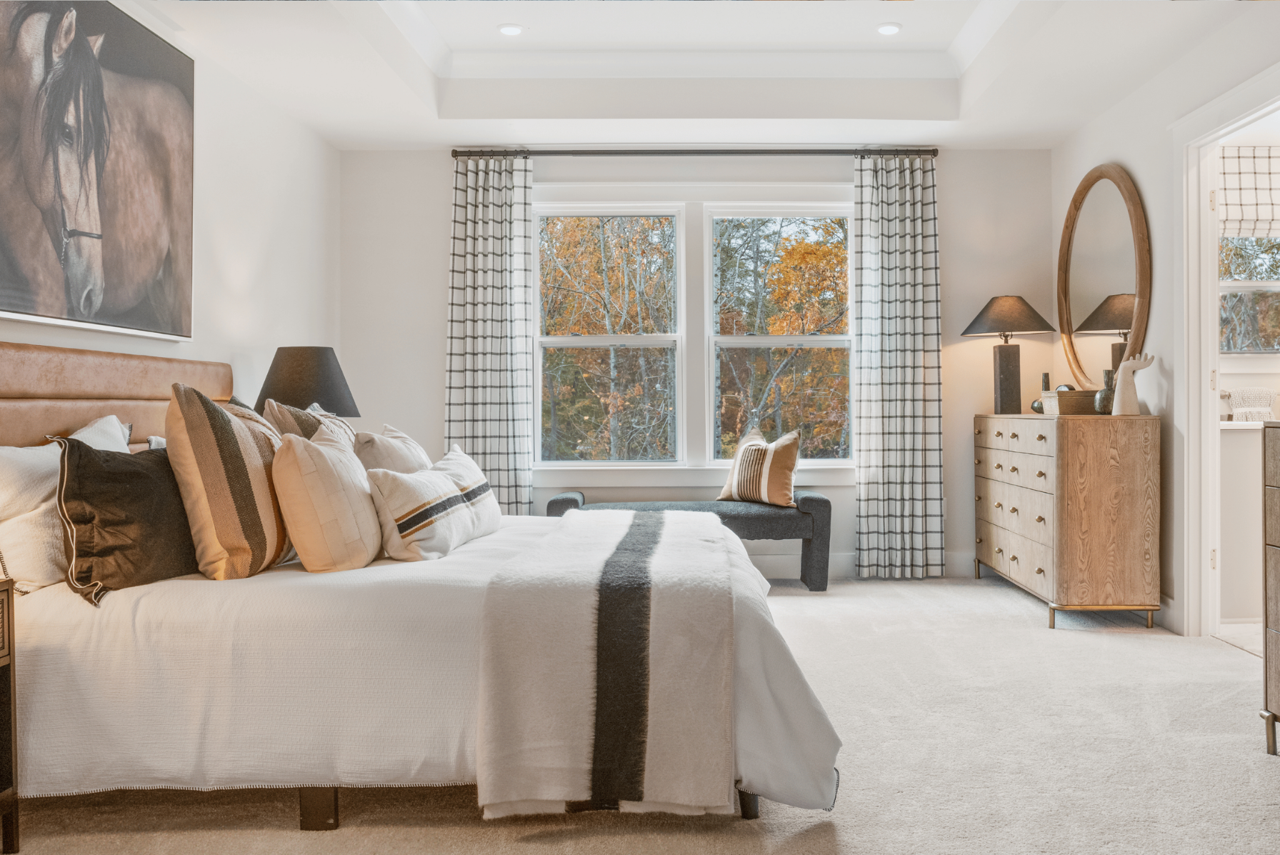 Modern bedroom with neutral tones, featuring a cozy bed, layered pillows, a horse painting, large windows, and elegant furniture.