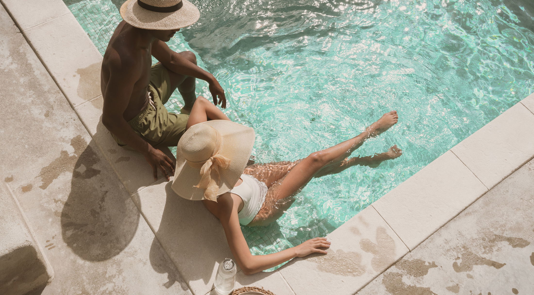 A couple relaxes by a sparkling turquoise pool, wearing sun hats and swimwear, enjoying a sunny day.