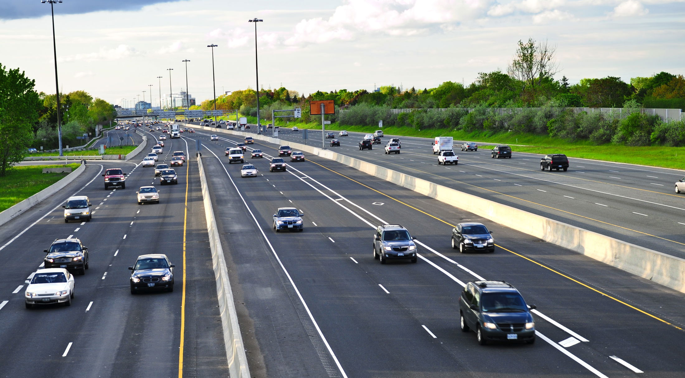 A multi-lane highway with light traffic, surrounded by green trees and under a partly cloudy sky.
