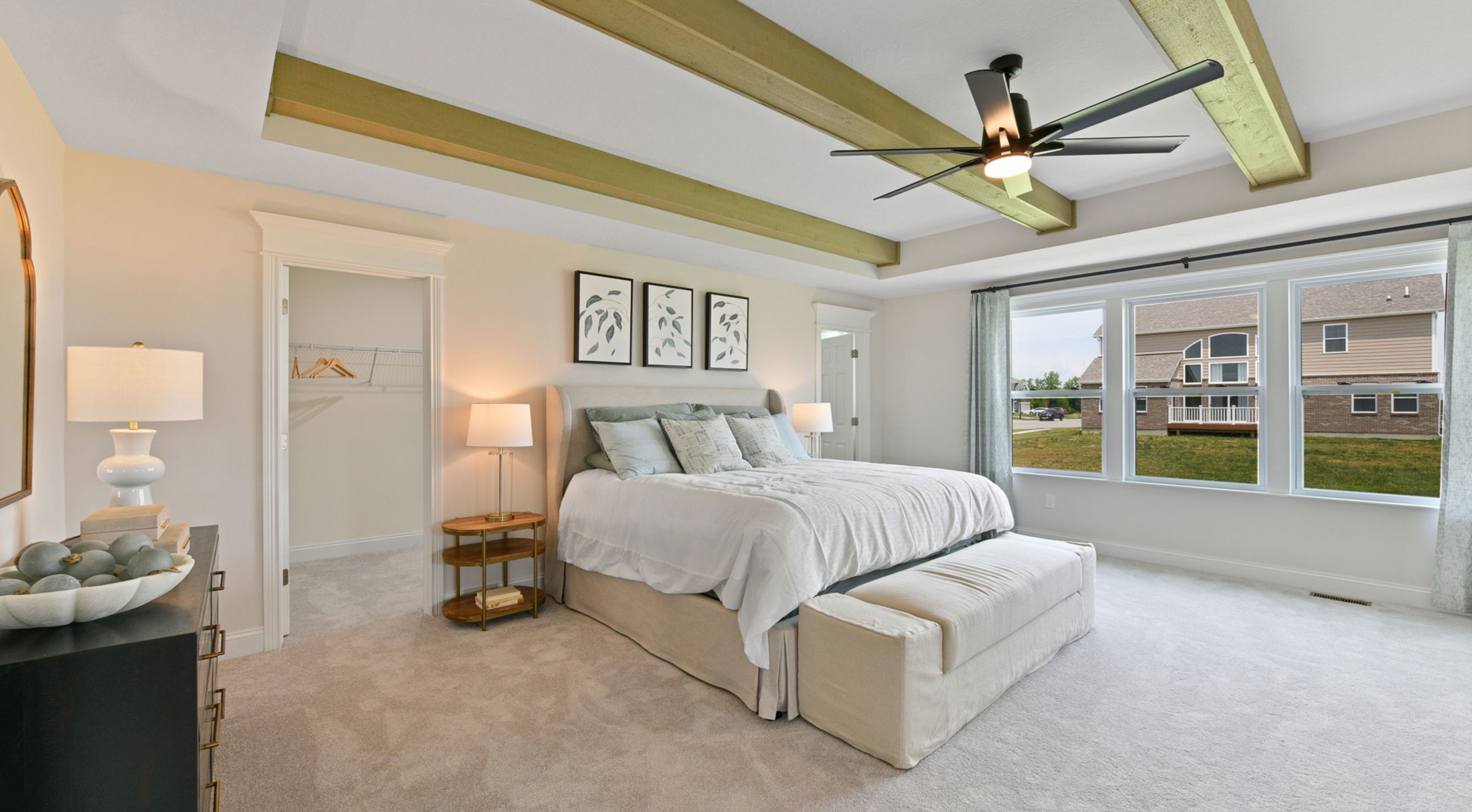 Modern bedroom with ceiling beams, stylish ceiling fan, and cozy decor featuring a well-lit space through large windows.