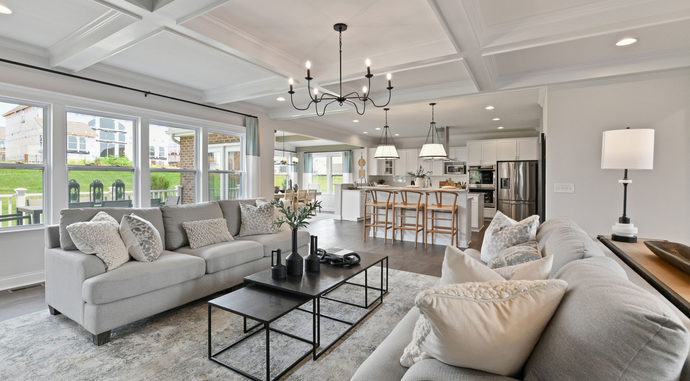 Modern open-concept living room with gray sofas, a coffered ceiling, contemporary chandelier, and spacious kitchen with island seating.