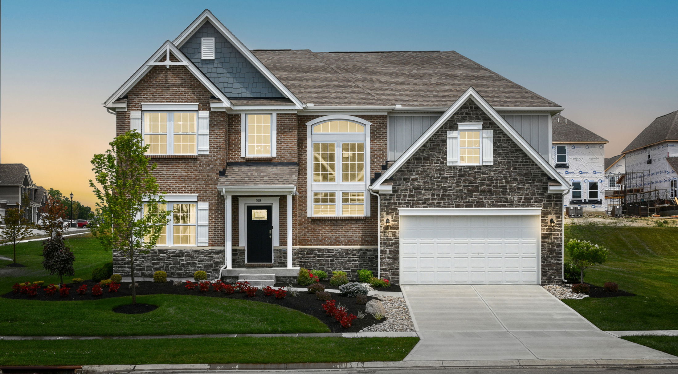 Modern two-story brick and stone house with a manicured front lawn and illuminated windows at sunset.