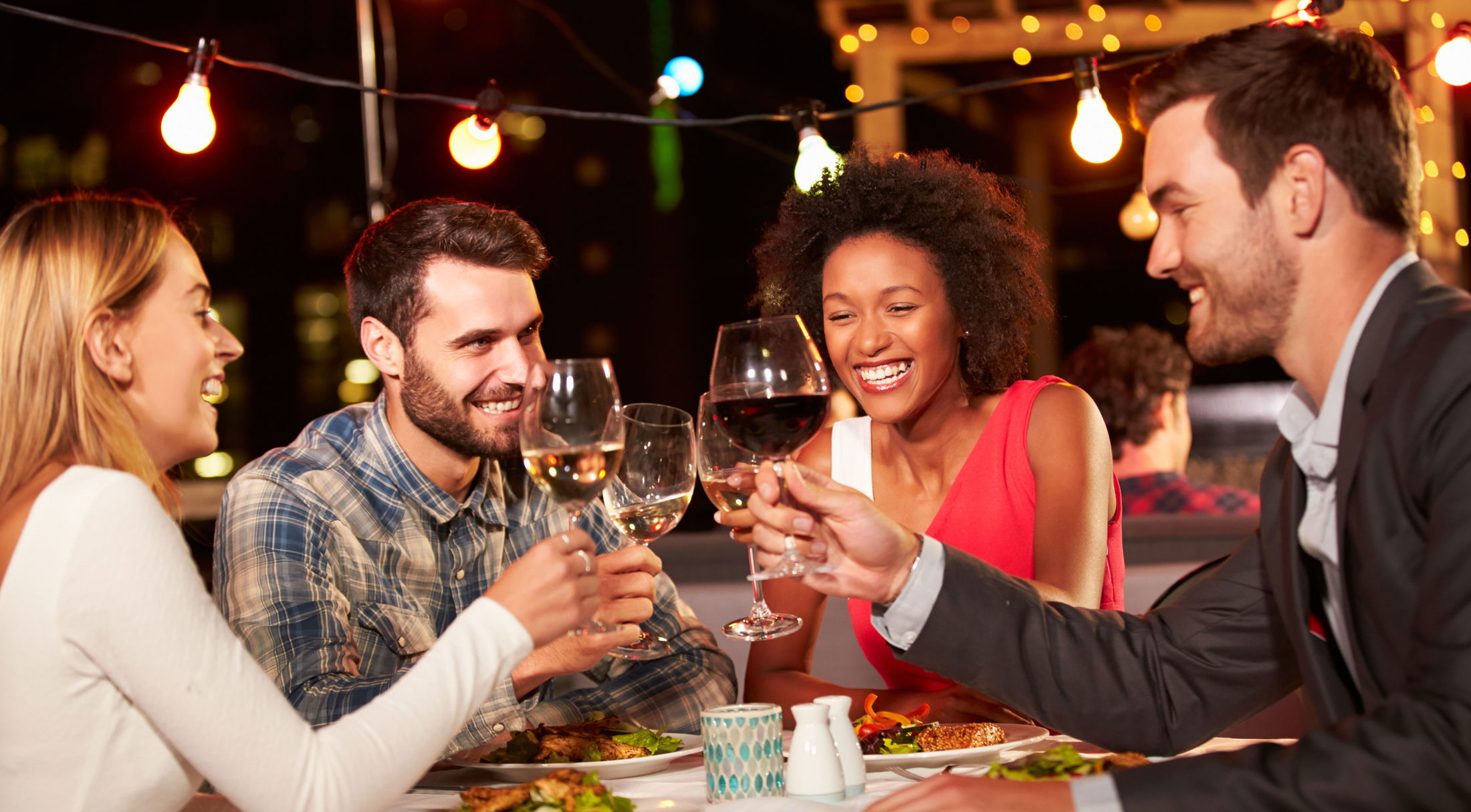 A group of friends enjoying a lively dinner with wine at an outdoor restaurant under string lights.