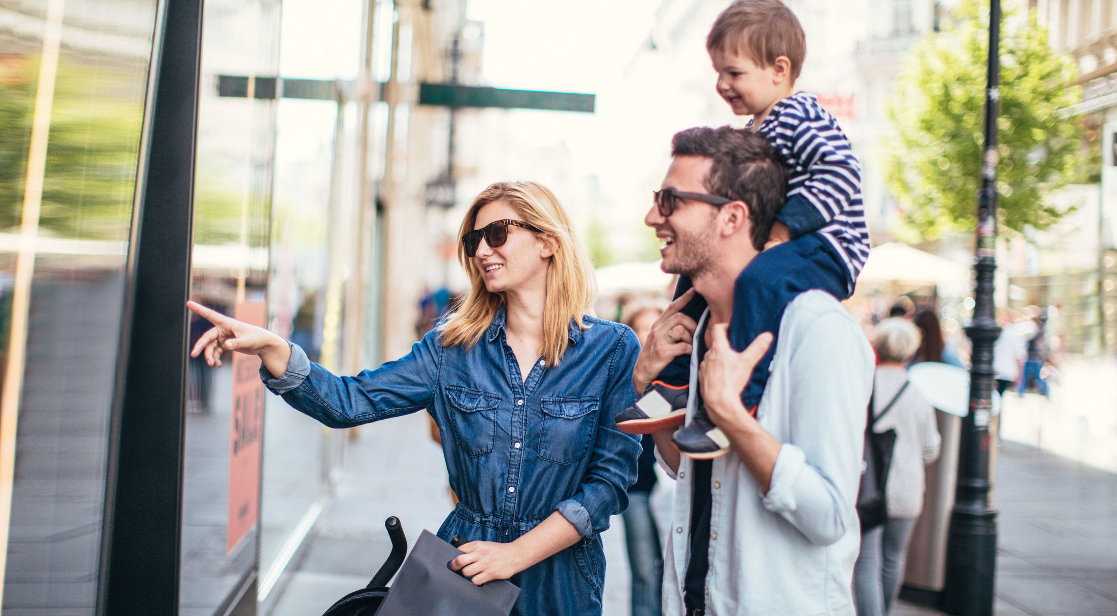 A family enjoys window shopping on a sunny day, with a child sitting on the father\\\'s shoulders and the mother pointing at a storefront.