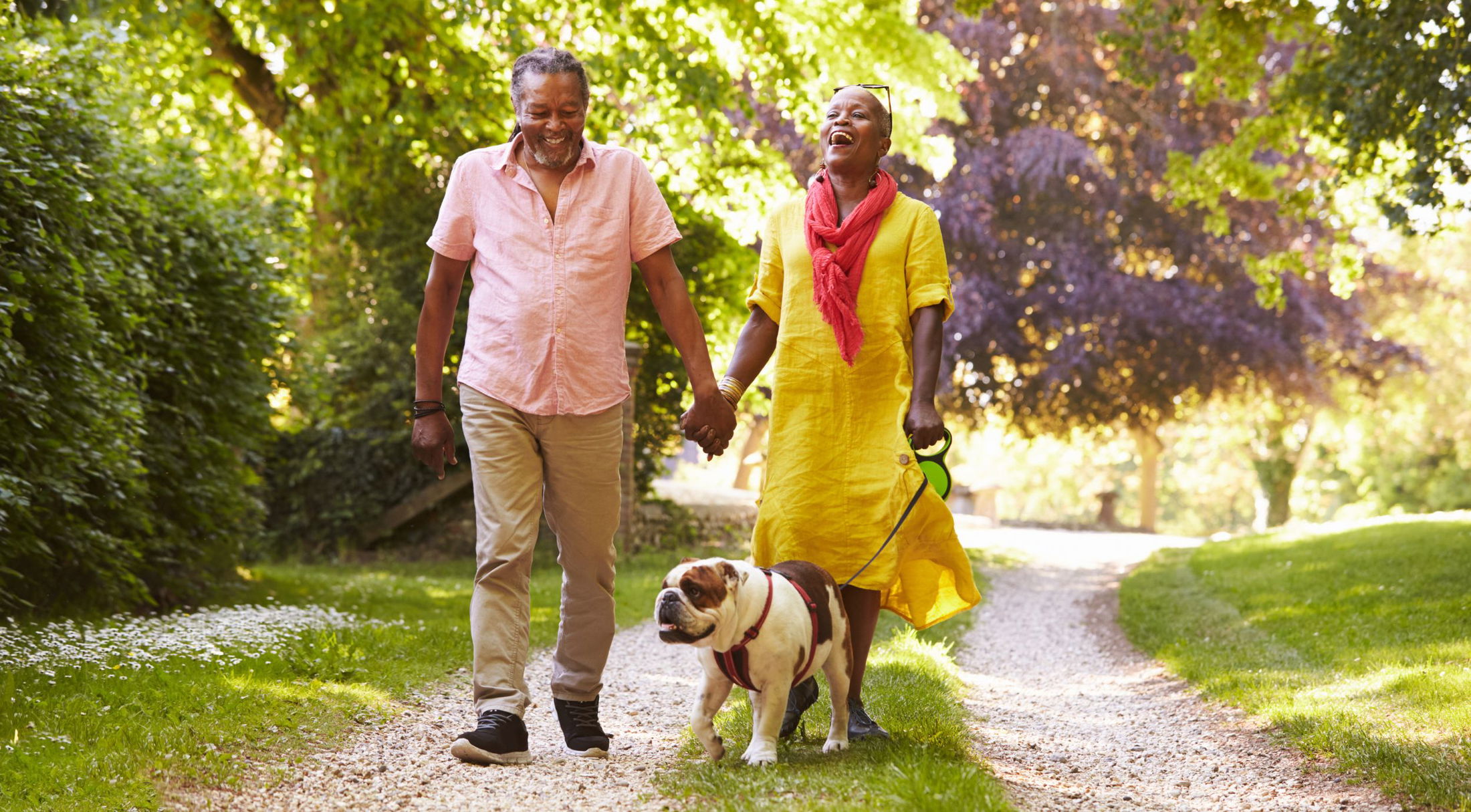 A joyful couple walks hand in hand with their bulldog along a scenic, tree-lined path.