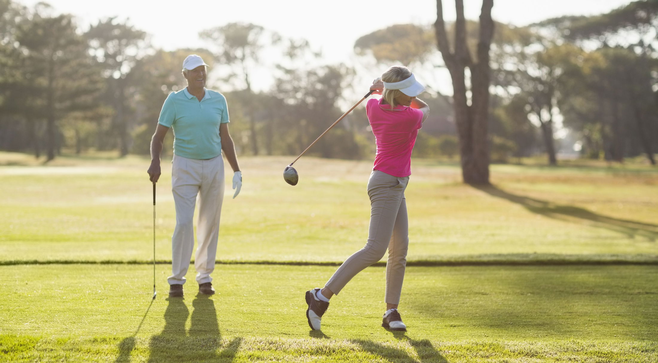 A woman in a pink shirt expertly swings a golf club on a sunny golf course, observed by a man in a blue shirt.