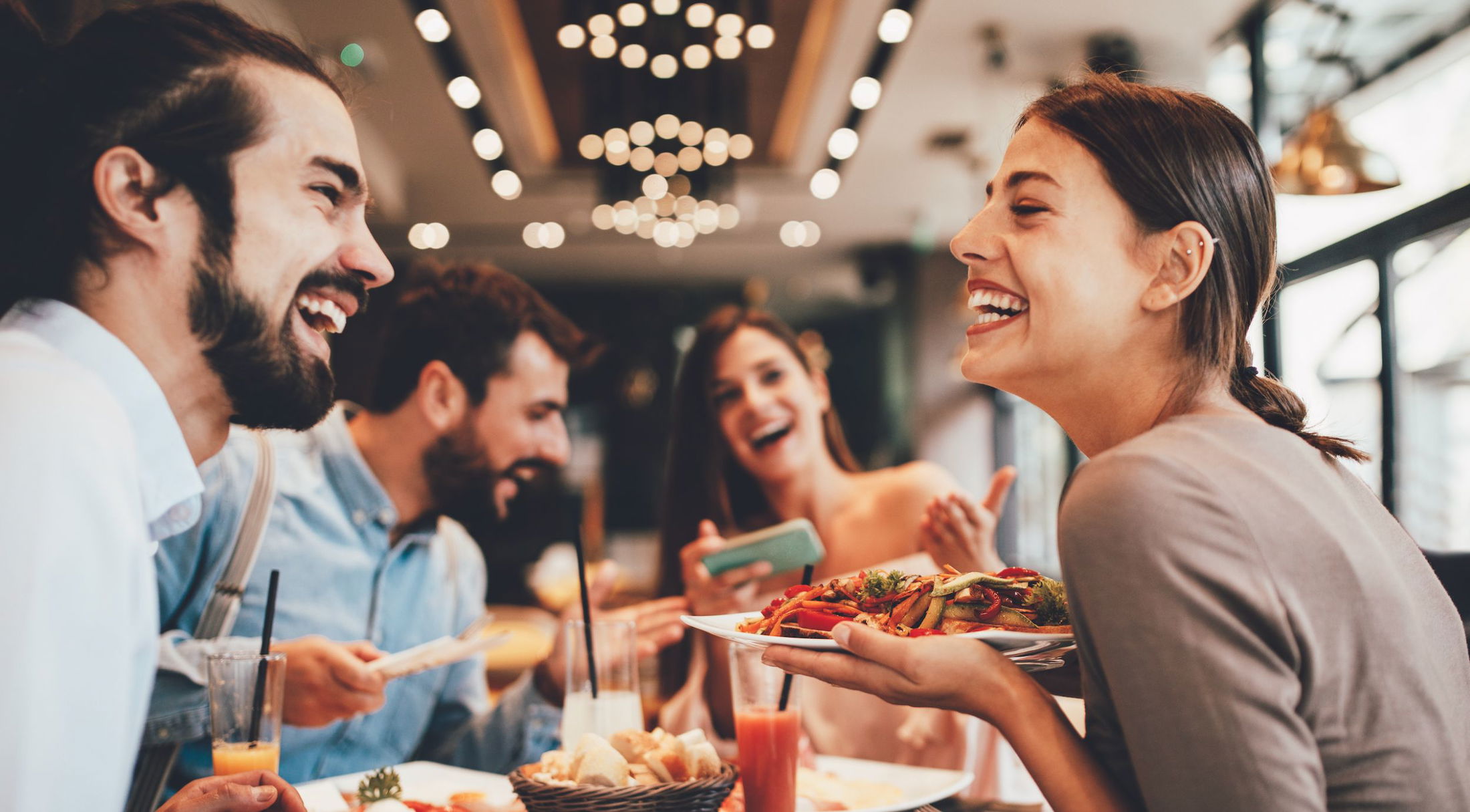 A group of friends laughing and enjoying a meal together at a trendy restaurant with warm lighting.