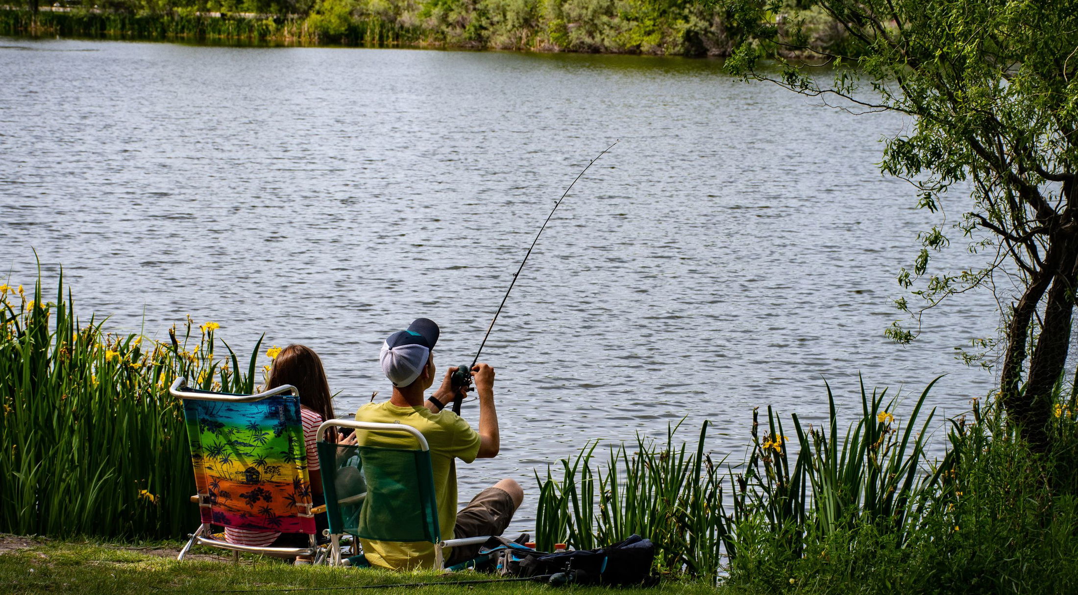 A man and woman relax in colorful chairs while fishing by a tranquil lakeside, surrounded by lush greenery.