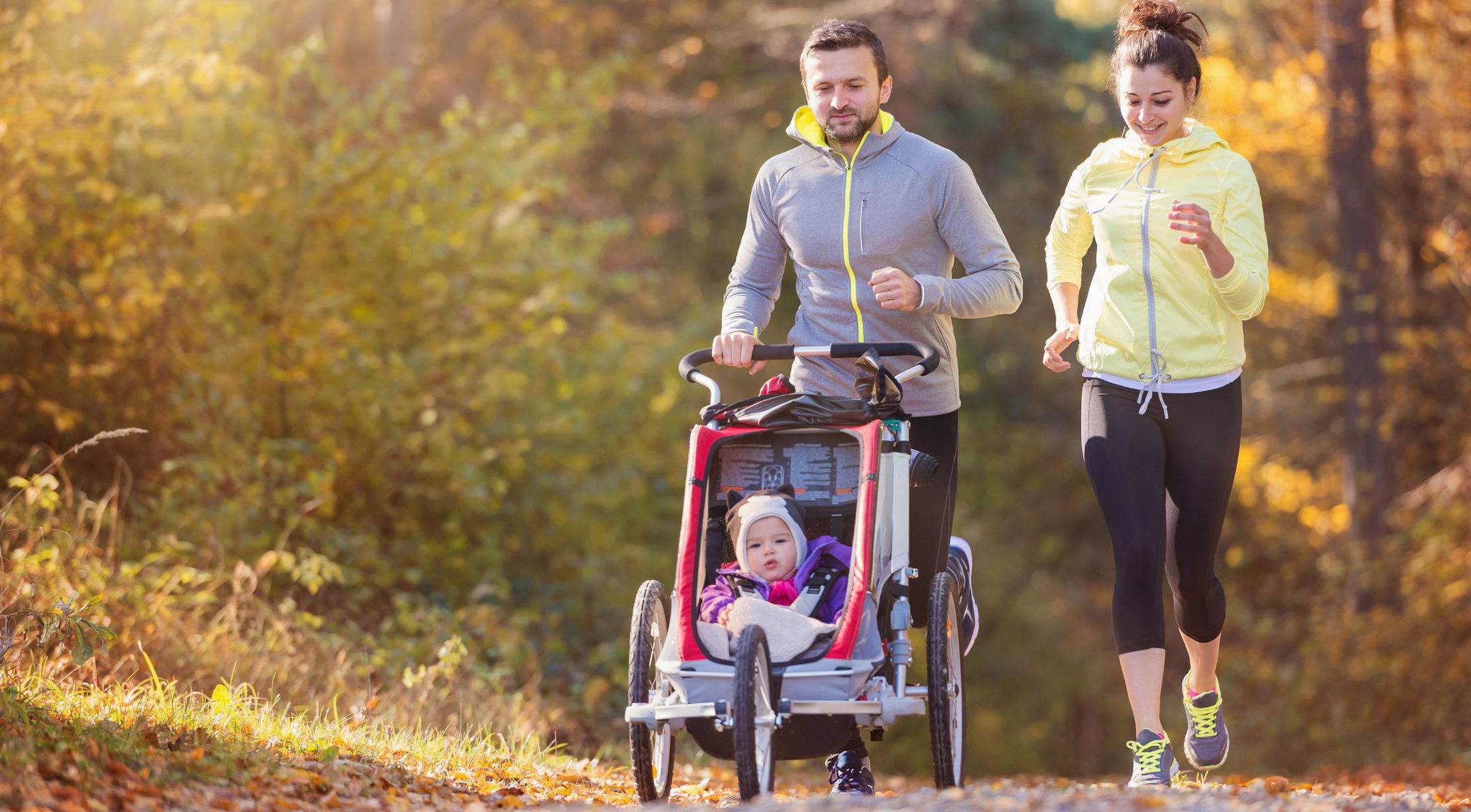 A couple jogging on a scenic autumn trail with a baby in a stroller, enjoying outdoor exercise.